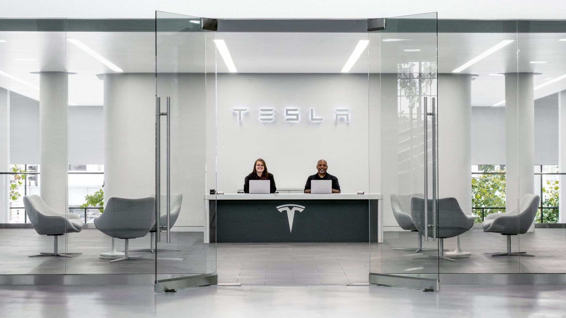 Tesla reception area with logo, two people behind a desk, glass doors, and seating.