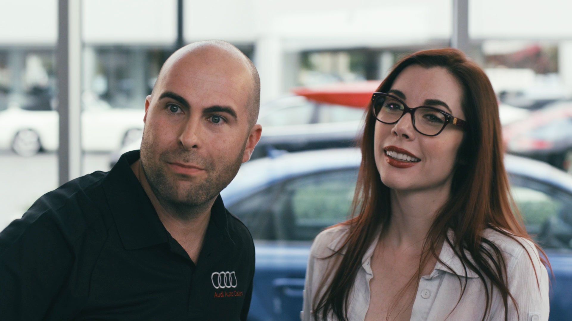 Man and woman in a car dealership, smiling. Man is bald. Woman wears glasses. Blue car and windows are in background.