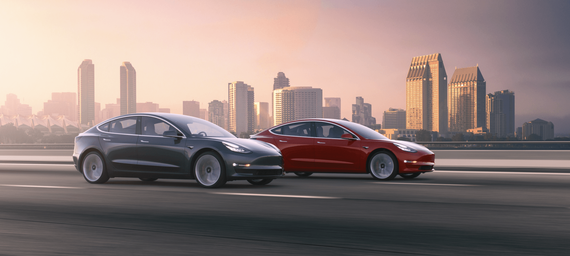 Two Tesla cars, grey and red, driving on a road with a cityscape in the background.