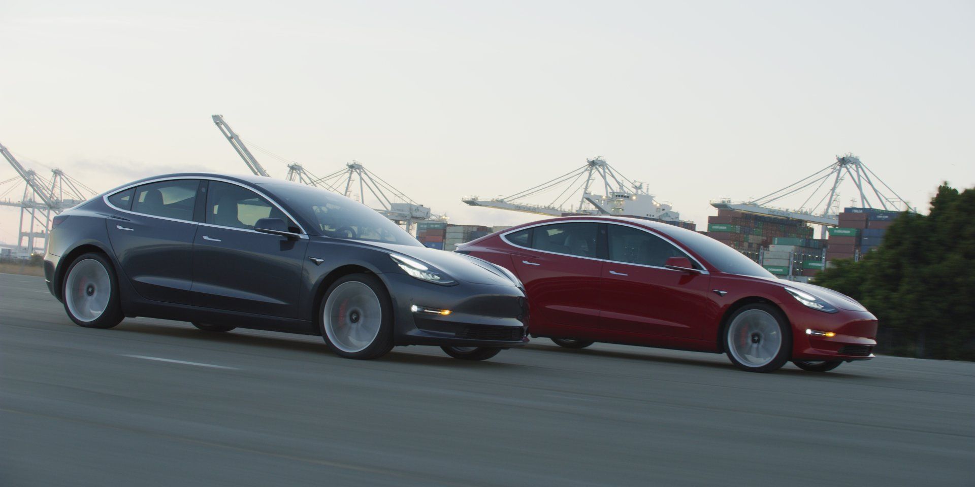 Two Tesla Model 3 cars, one gray and one red, driving on a road with industrial cranes in the background.