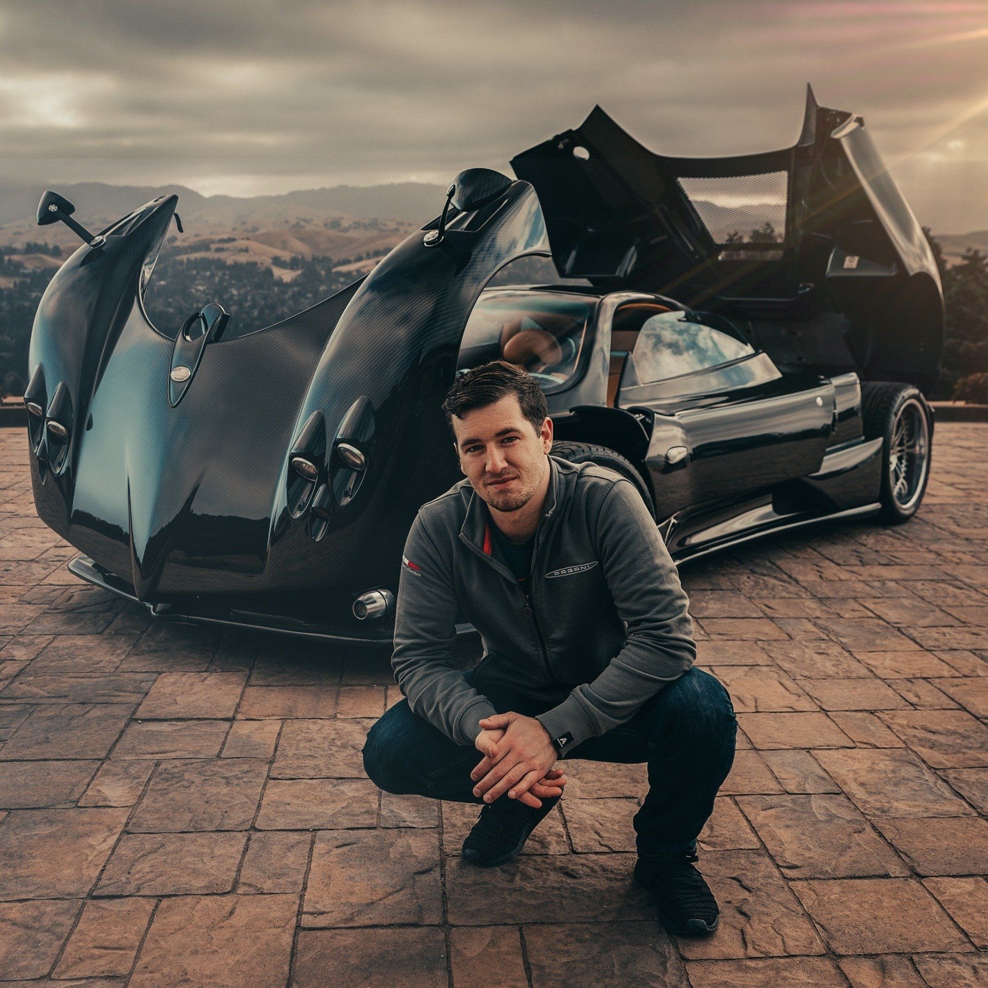 Man crouching in front of a black Pagani Zonda. He wears a gray jacket and jeans. Outdoors, mountain backdrop.