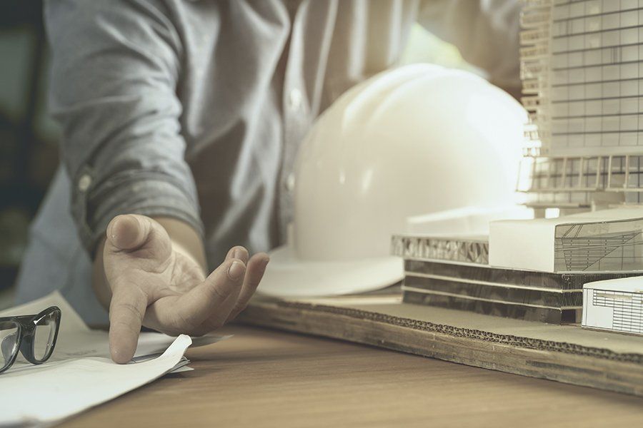 Arquitecto con maqueta de edificio y casco, haciendo un gesto hacia un plano de diseño sobre una mesa de madera.