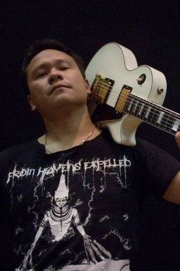 Persoon poseert met een witte elektrische gitaar, gekleed in een zwart T-shirt met print, tegen een donkere achtergrond.