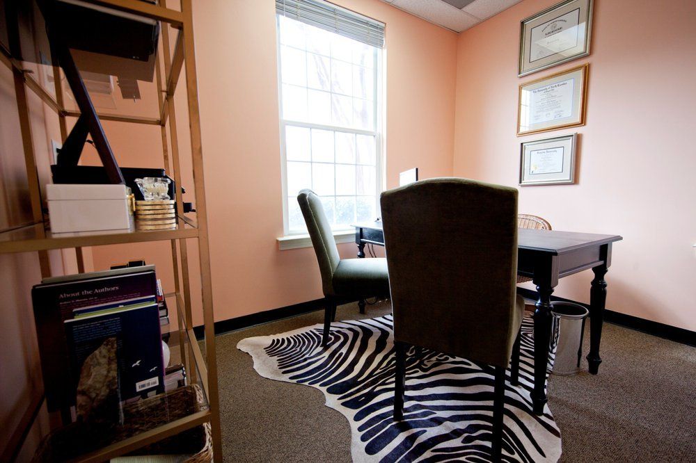 Small office with desk, chairs, window, shelves, and framed certificates on peach walls.