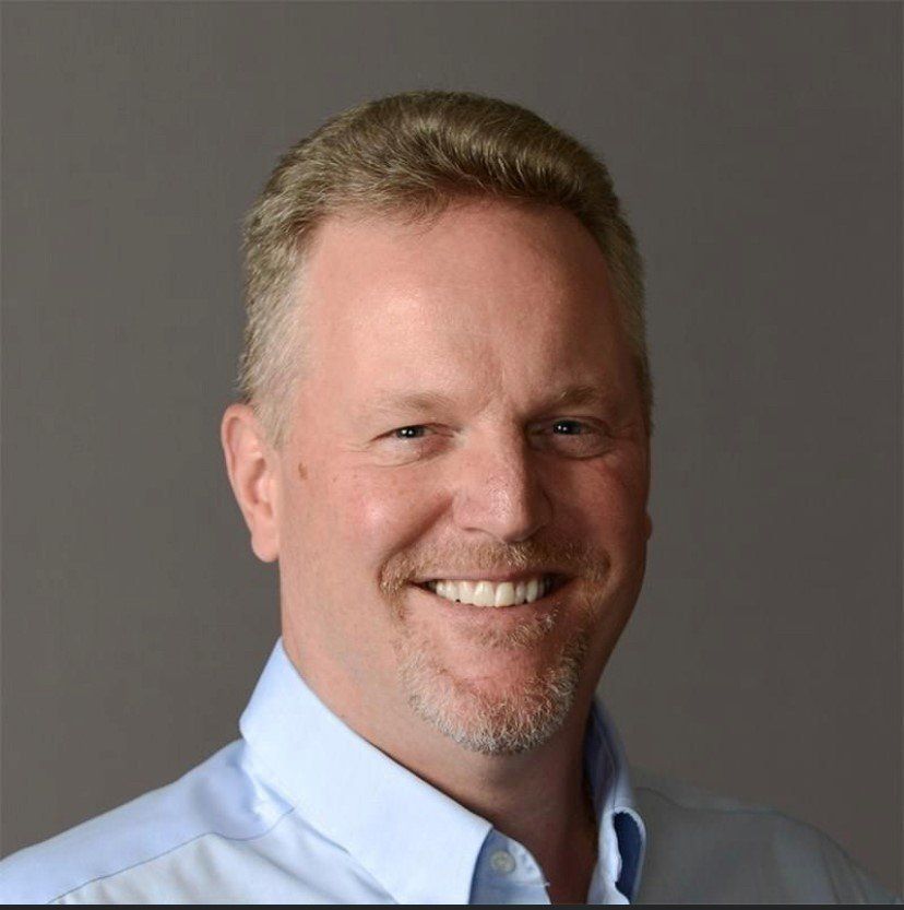 Man with fair skin, smiling, wearing a light blue button-down shirt.