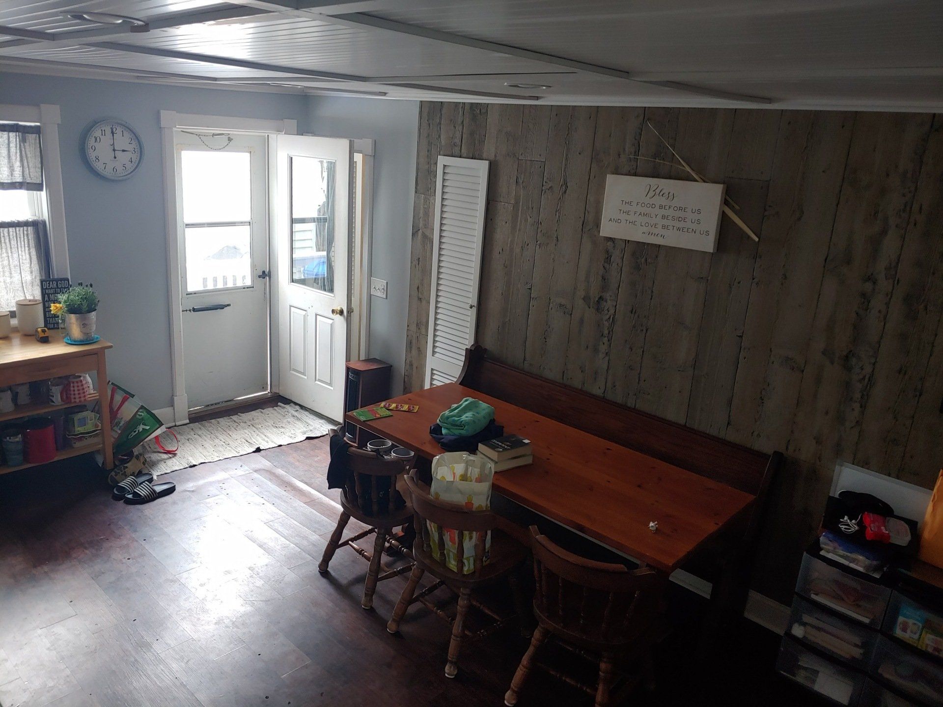 Small cluttered room with wooden table, chairs, open door, wall clock, and shelves.
