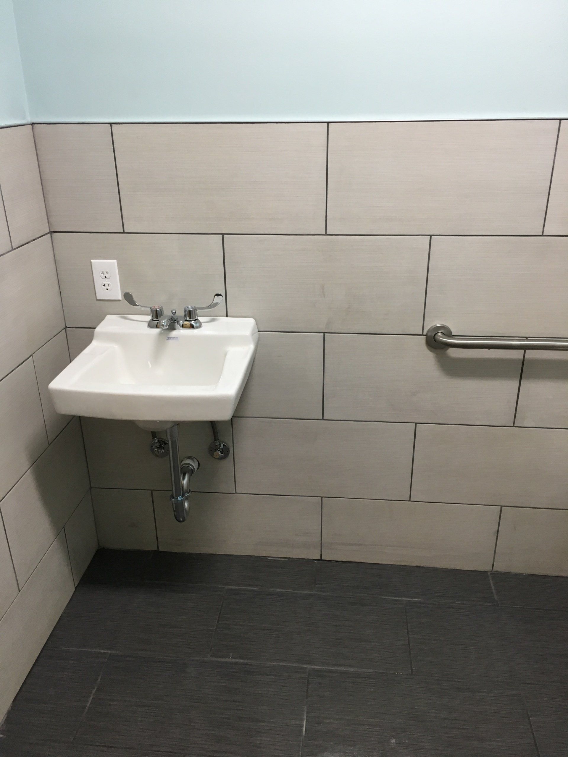 Small accessible restroom sink with grab bar, tiled walls, and dark floor.