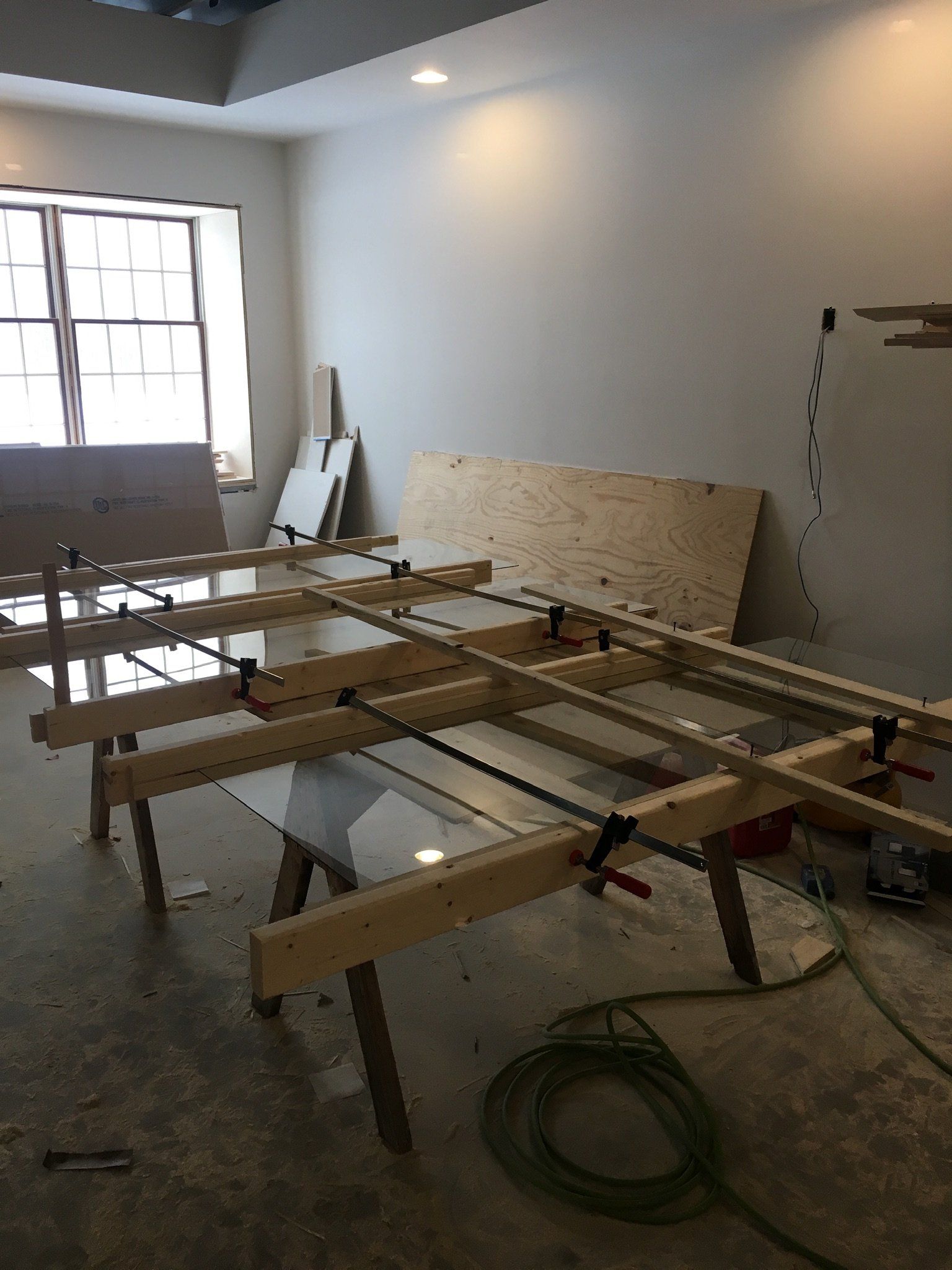 Empty workshop with a wooden workbench frame, plywood sheets, and a coiled green hose near a window.