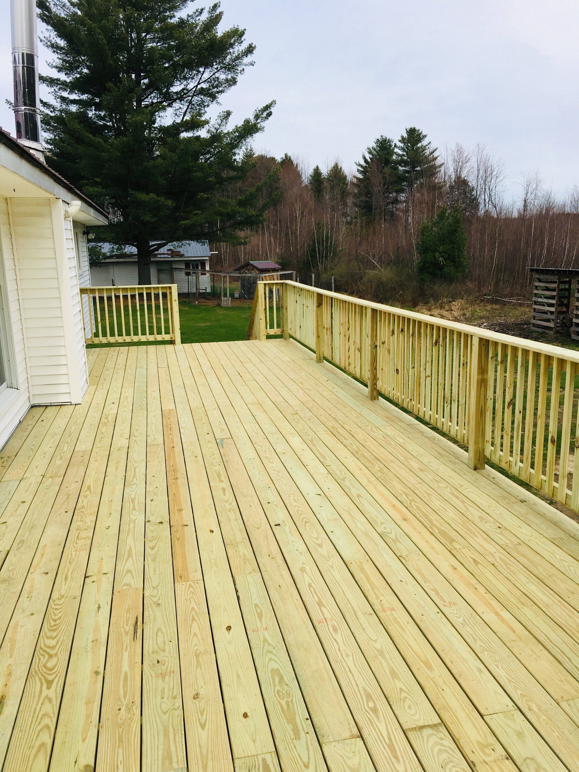 Freshly built wooden deck beside a white house, with railing and a wooded yard view