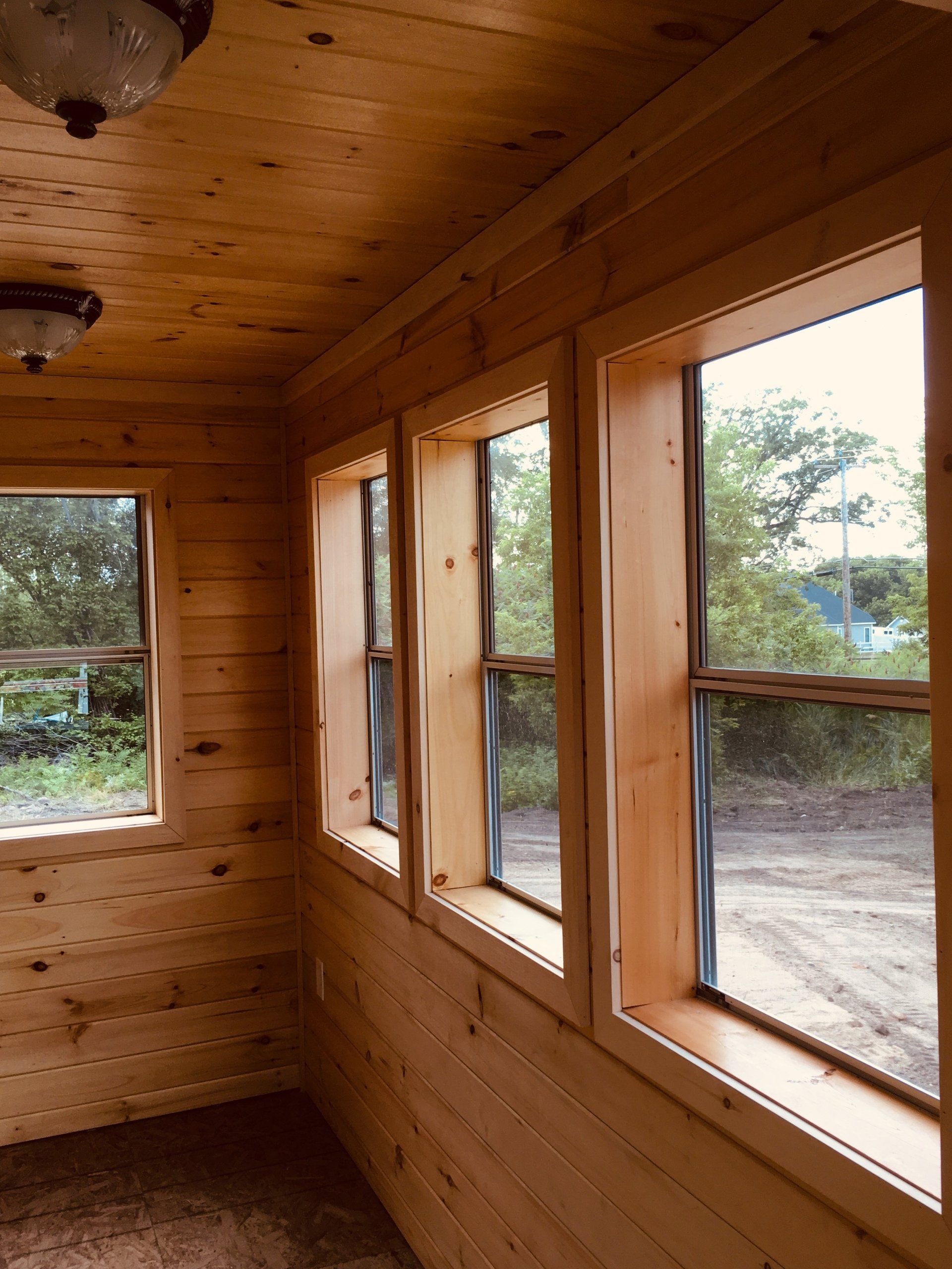 Wood-paneled cabin interior with three tall windows overlooking trees and dirt outside.