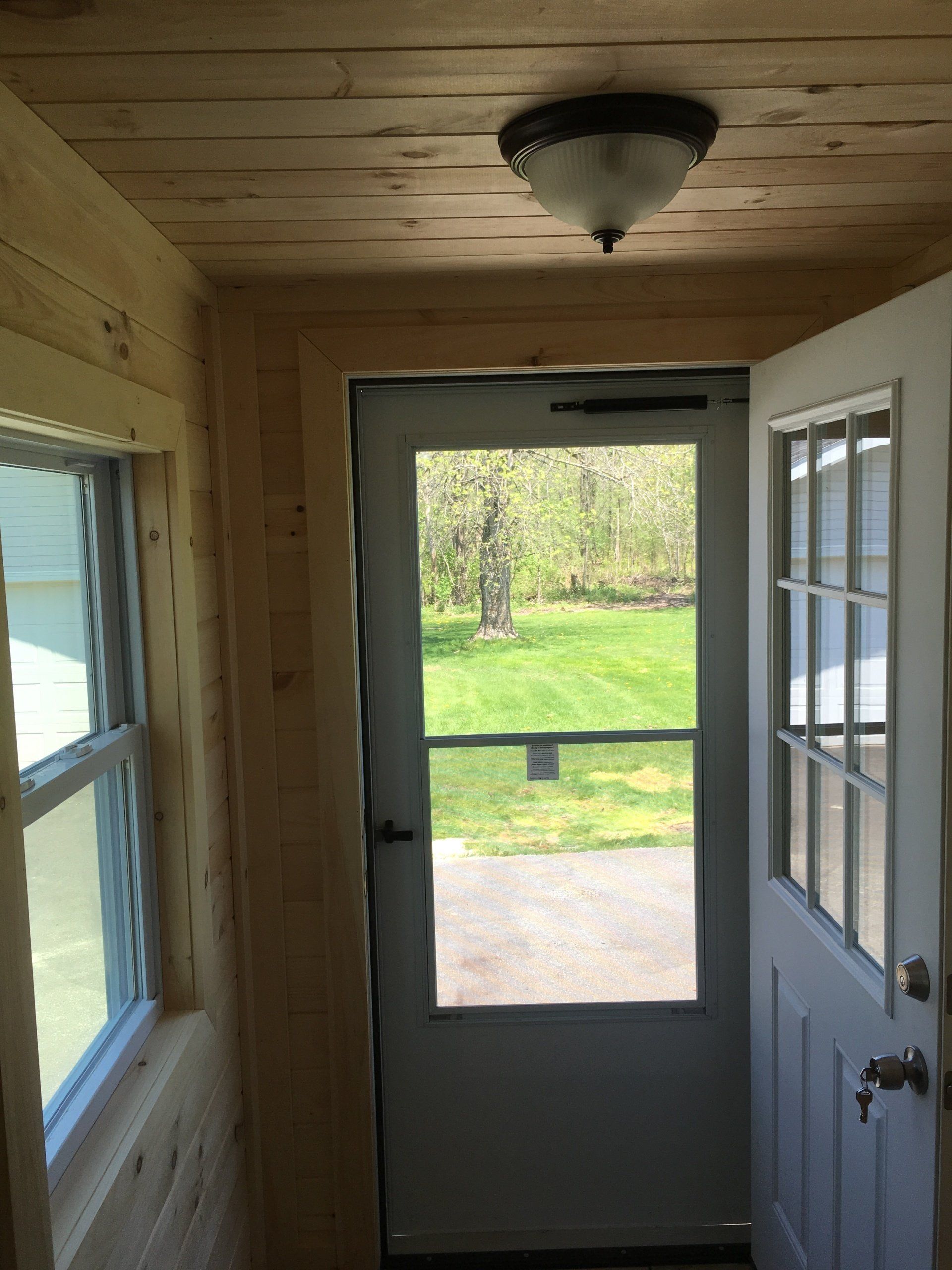 Narrow porch with wood-paneled walls, ceiling light, and open door looking out to a sunny yard.