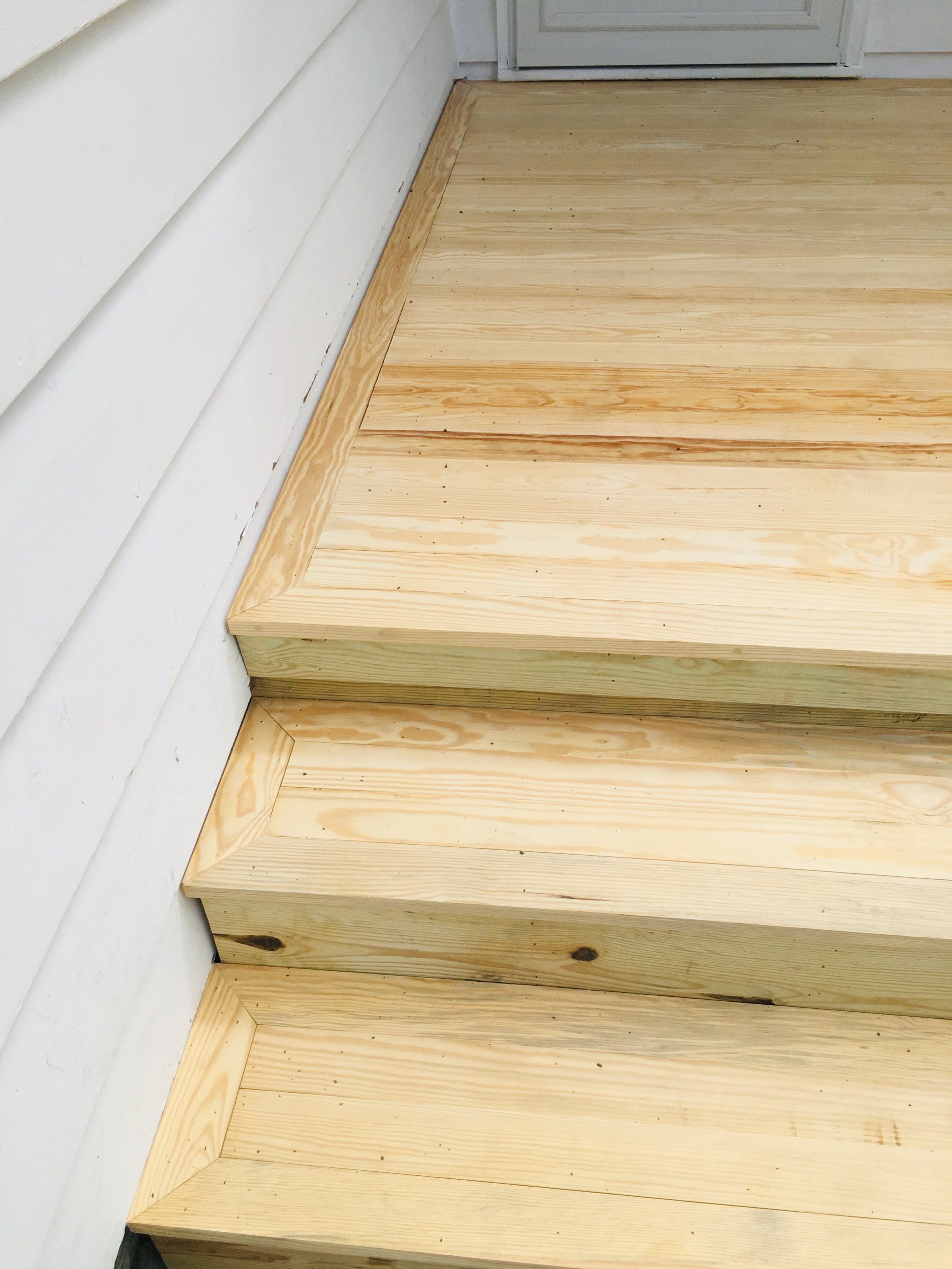Bare wooden porch steps beside a white exterior wall and closed door