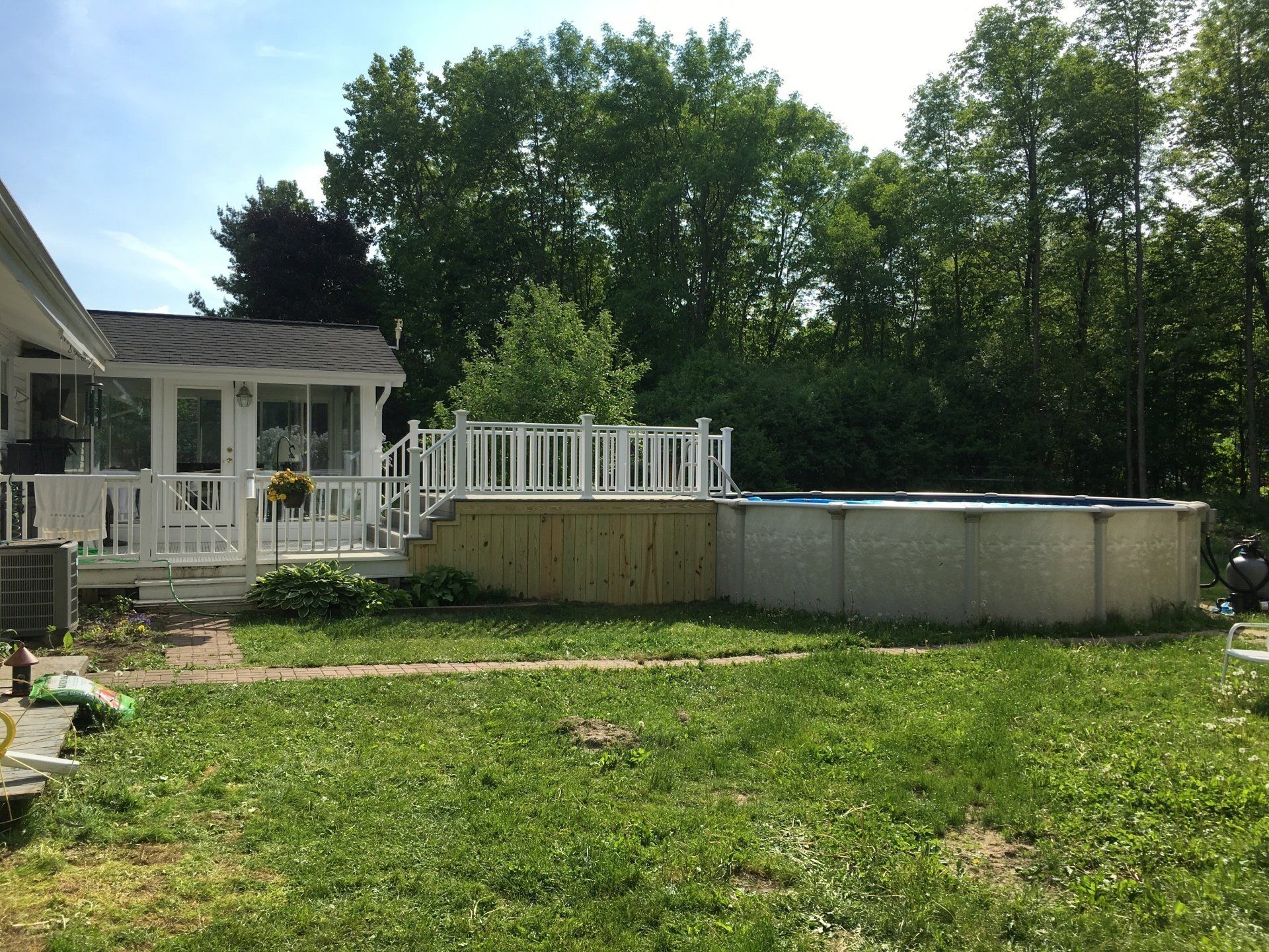 Backyard with a deck, above-ground pool, and green lawn beside a white house and trees