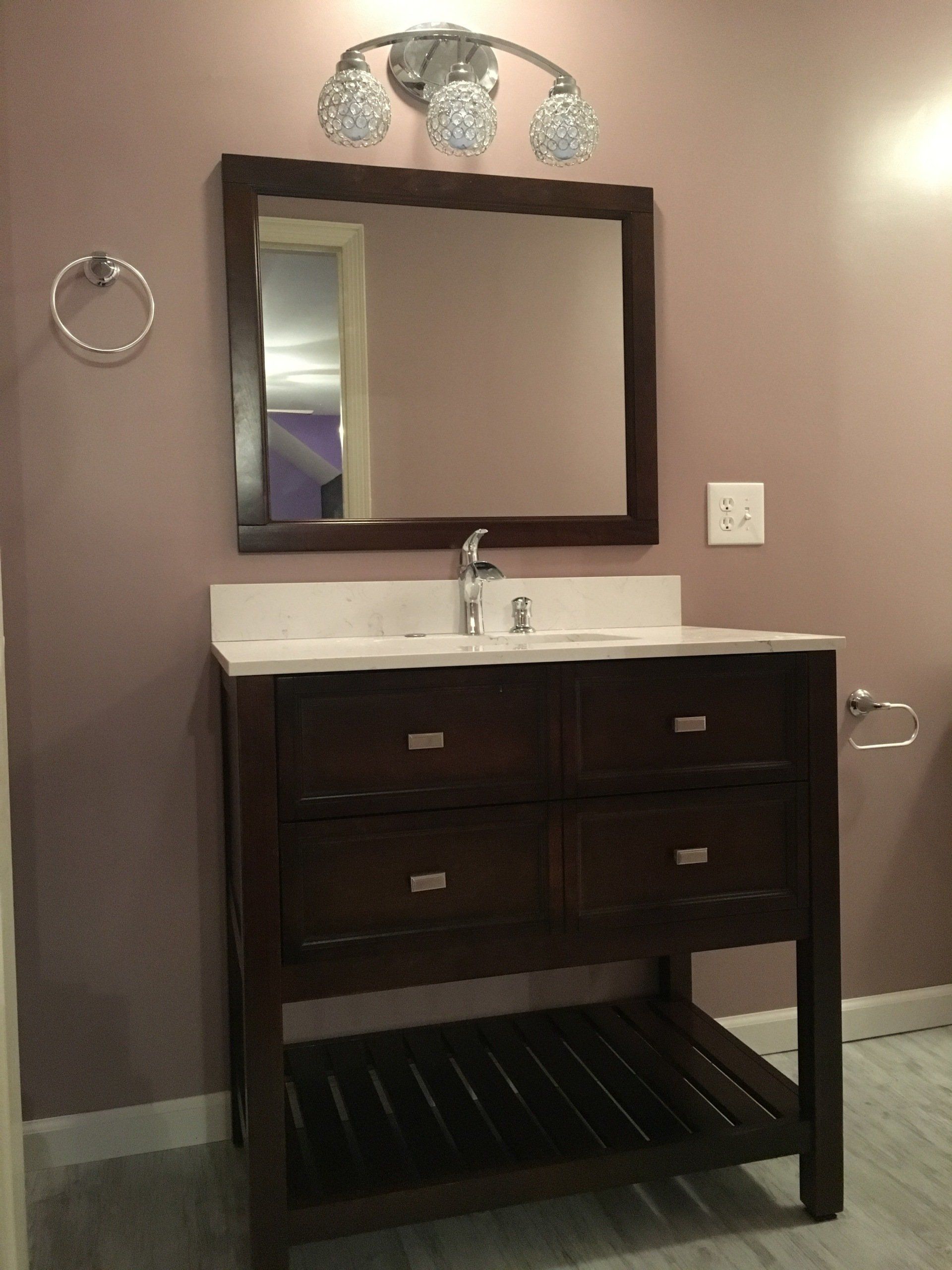 Dark wood bathroom vanity with white sink, mirror, wall sconce, and towel ring on gray walls.