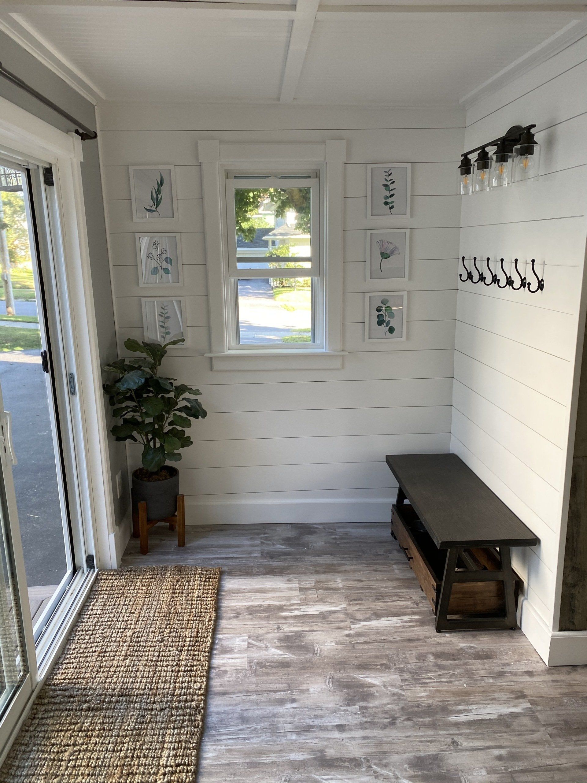 Bright entryway with white walls, framed art, a bench, rug, and a potted plant by the door.