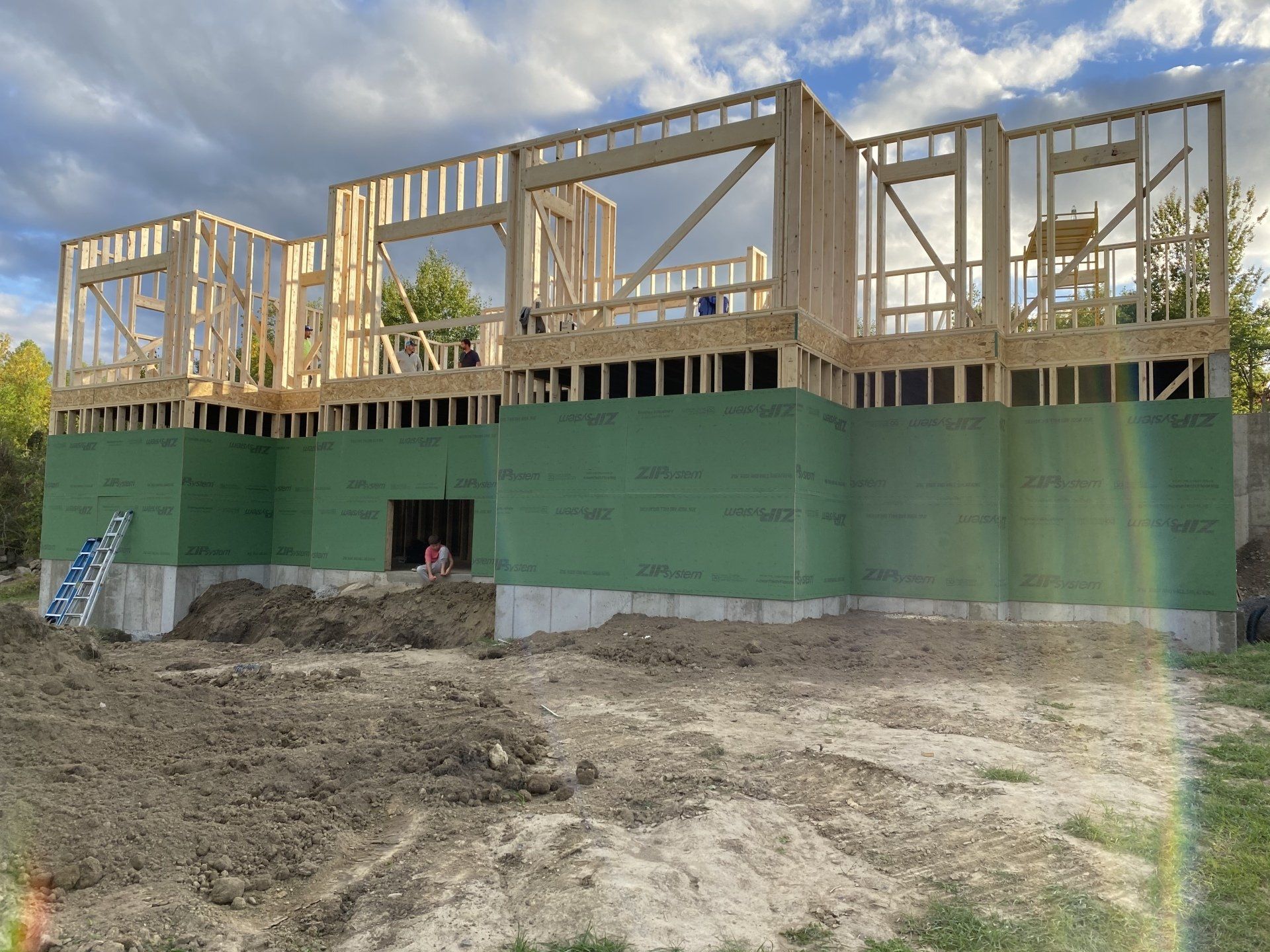 Partially built house with exposed wooden framing and green wrapped lower walls on a dirt lot.