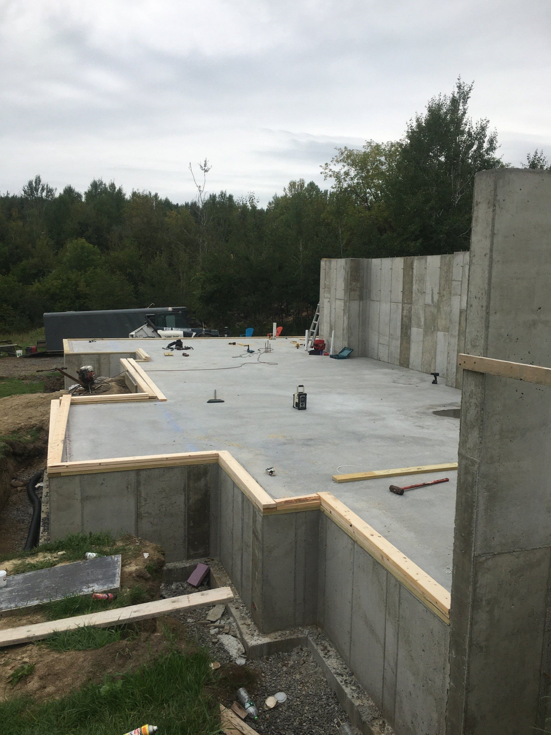 Concrete foundation slab and retaining walls under construction beside a wooded area, with tools scattered on top.