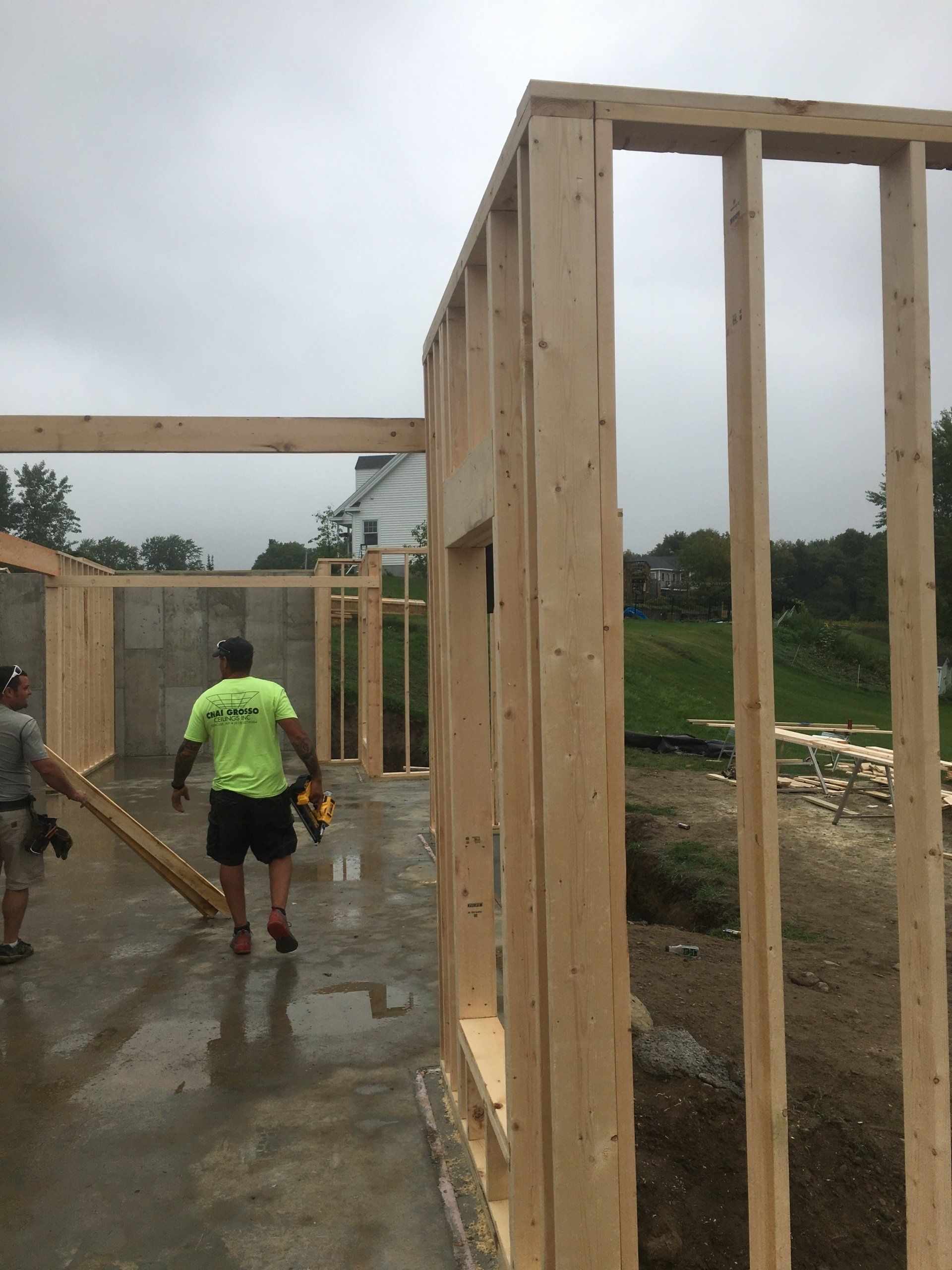 Wooden building frames under construction on a wet concrete slab, with two workers walking through.