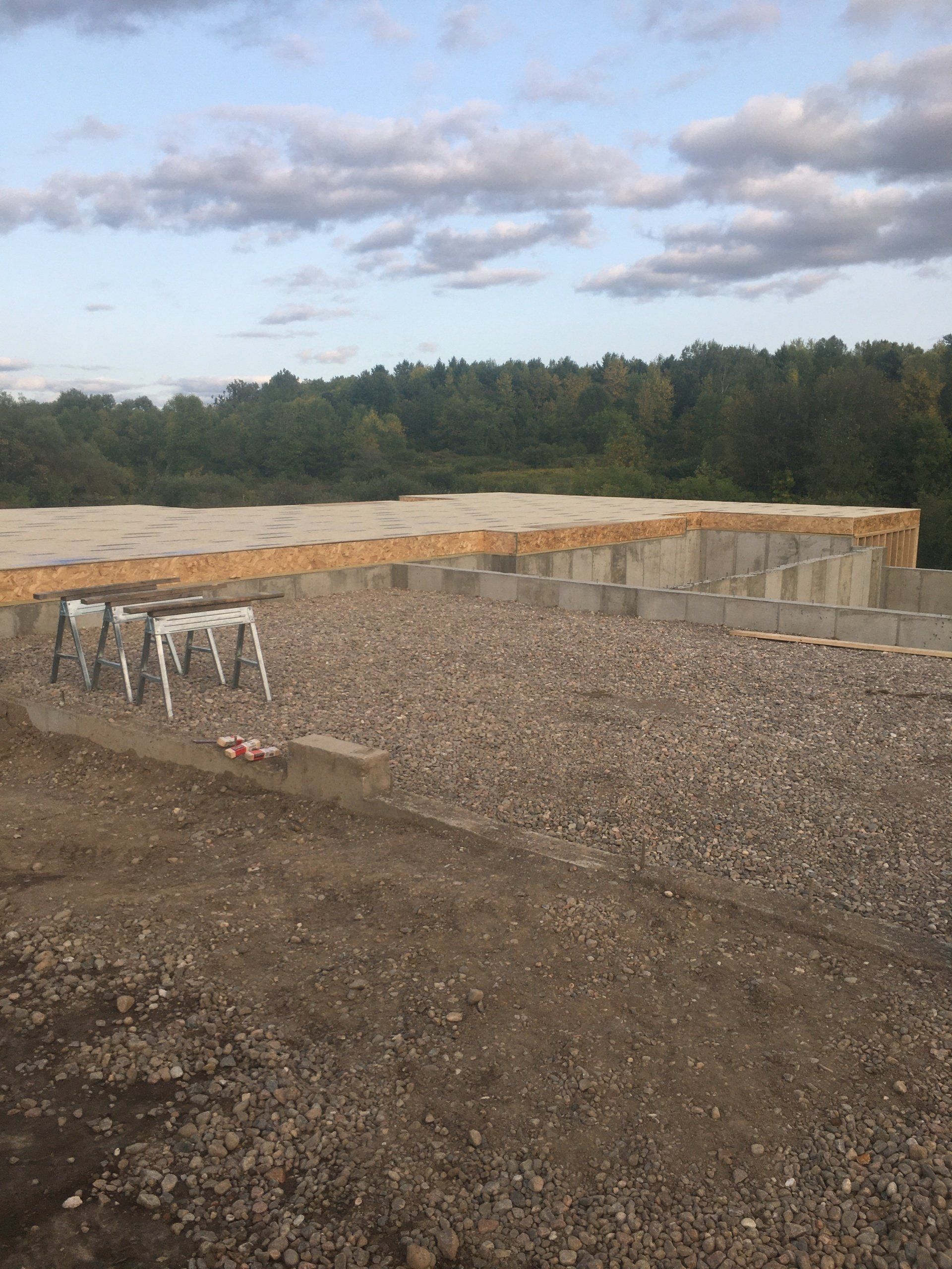 Gravel foundation site with concrete retaining wall and wooden forms under a cloudy sky.