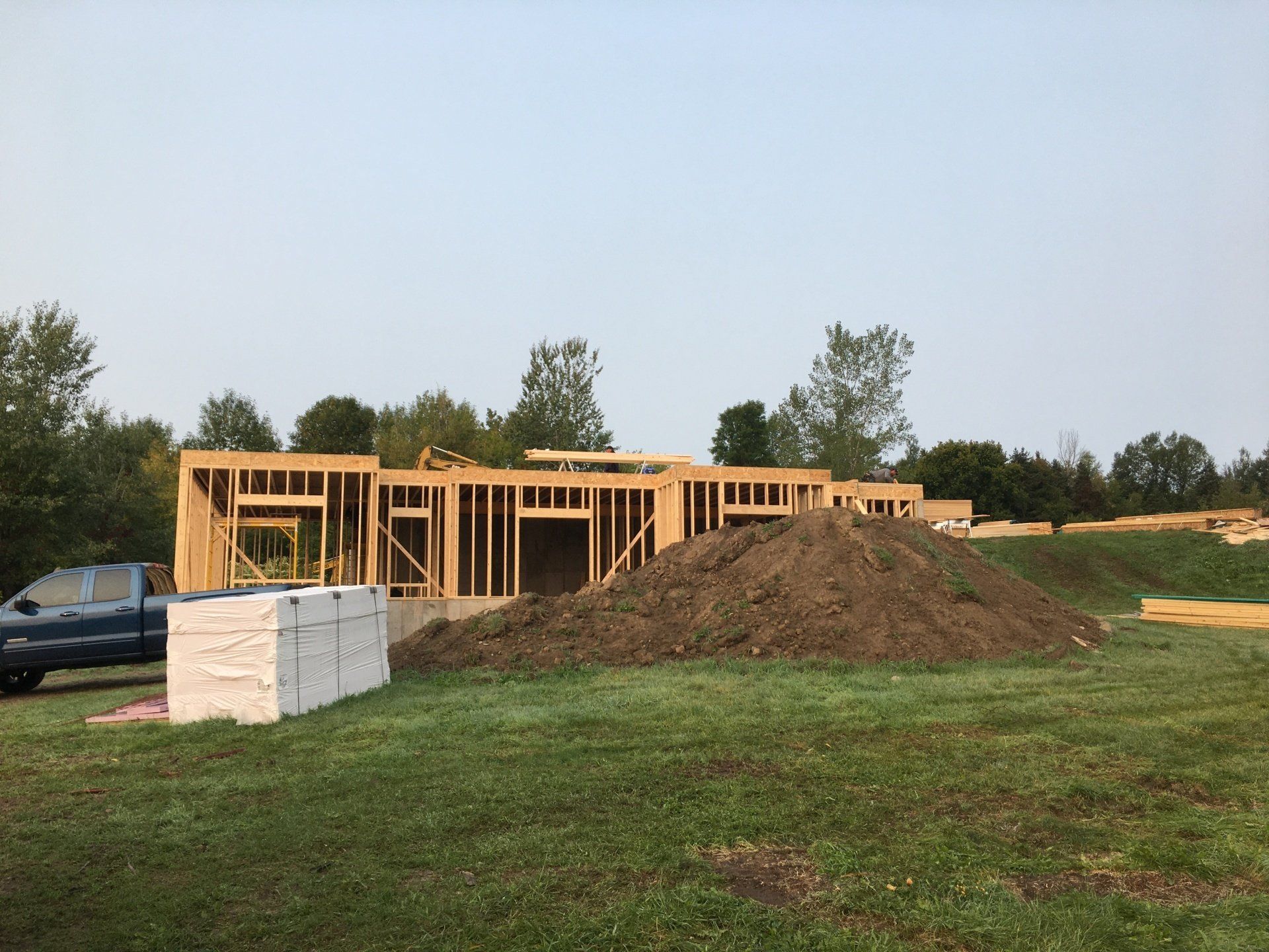 Partially built wooden house with a large dirt mound in front, on a grassy lot under a clear sky