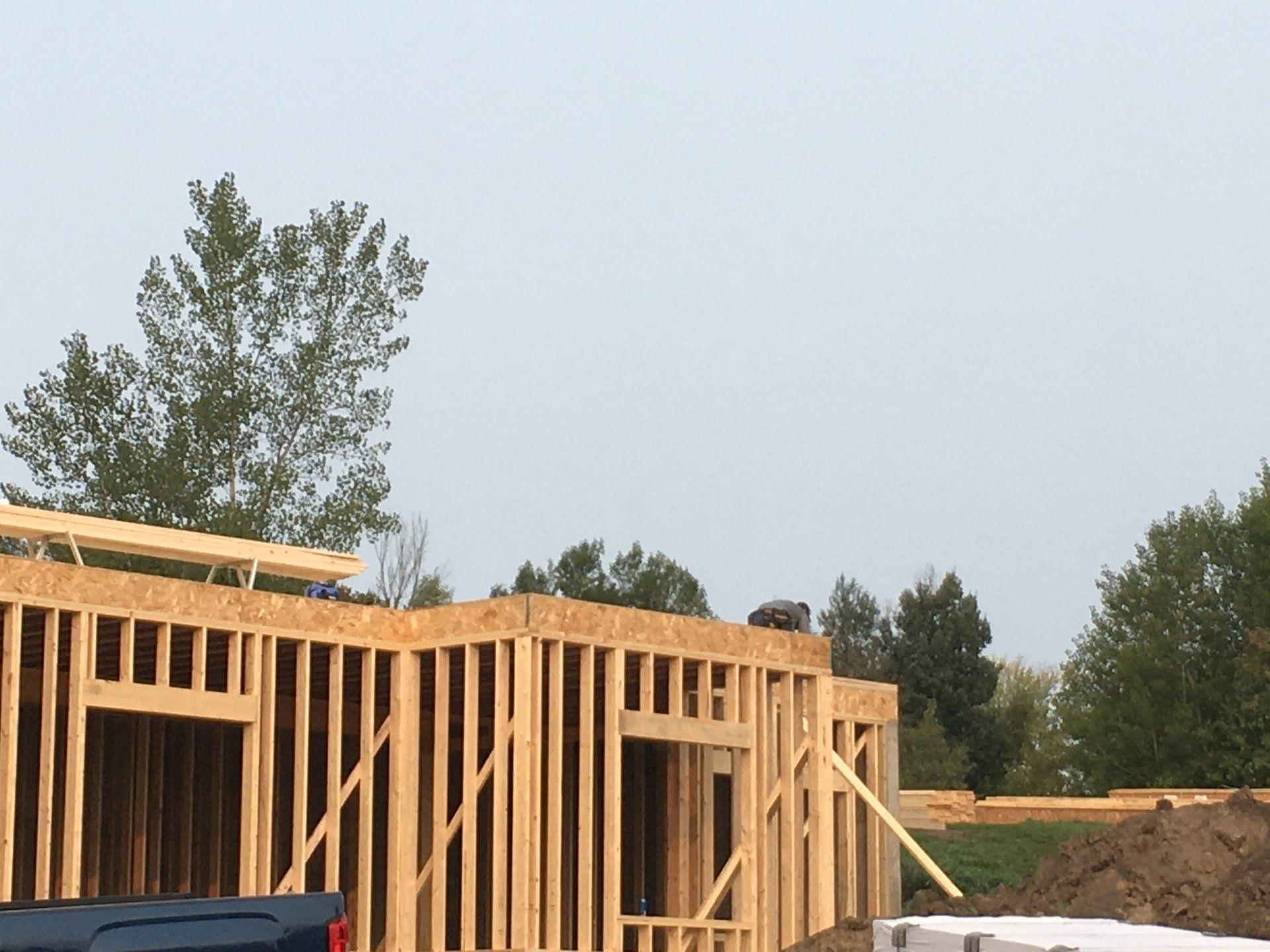 Wood-frame house under construction with vertical studs against a pale sky and trees in the background