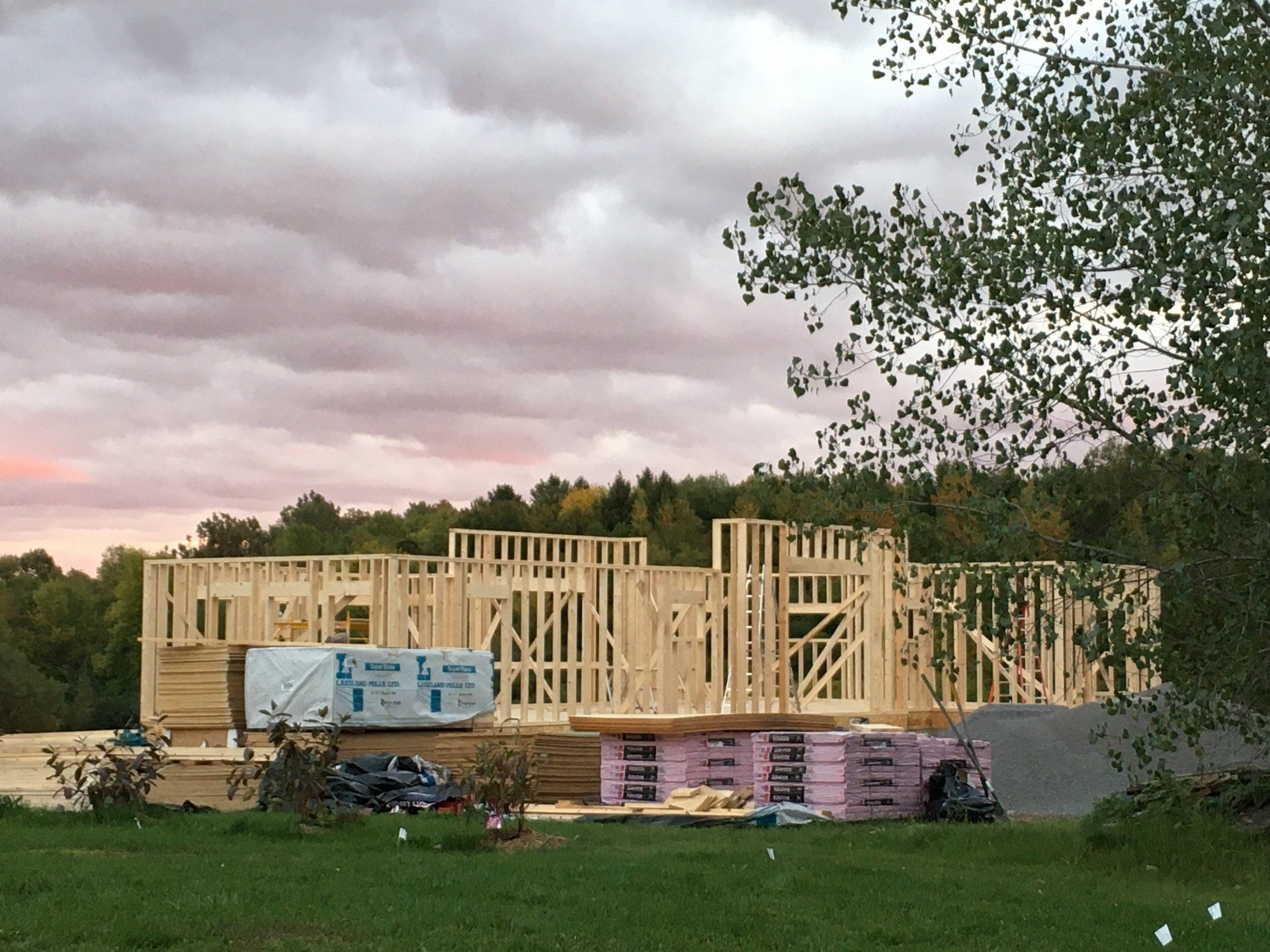 Partially built wooden house frame on a construction site with lumber piles and cloudy sky