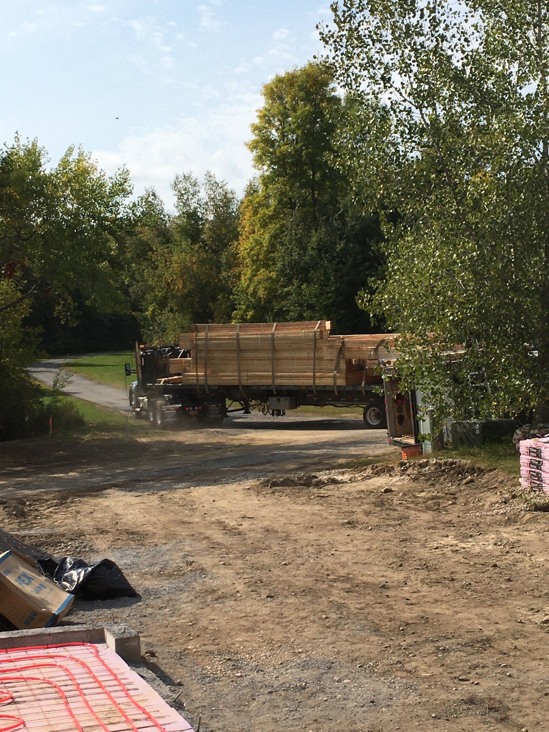 Flatbed truck on a dirt road in a wooded area, with stacked wood and a partly visible tarp in the foreground