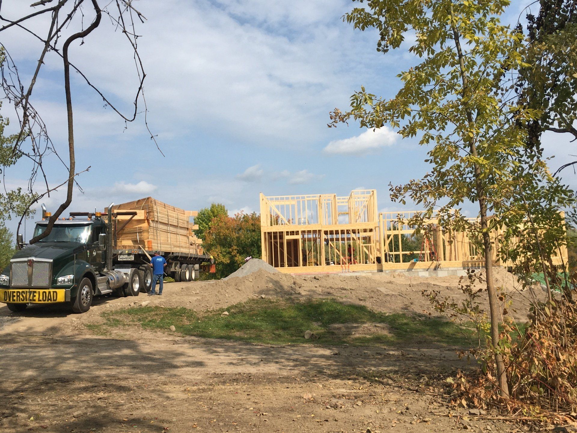 House under construction with a lumber truck, workers, and wooden framing on a dirt lot