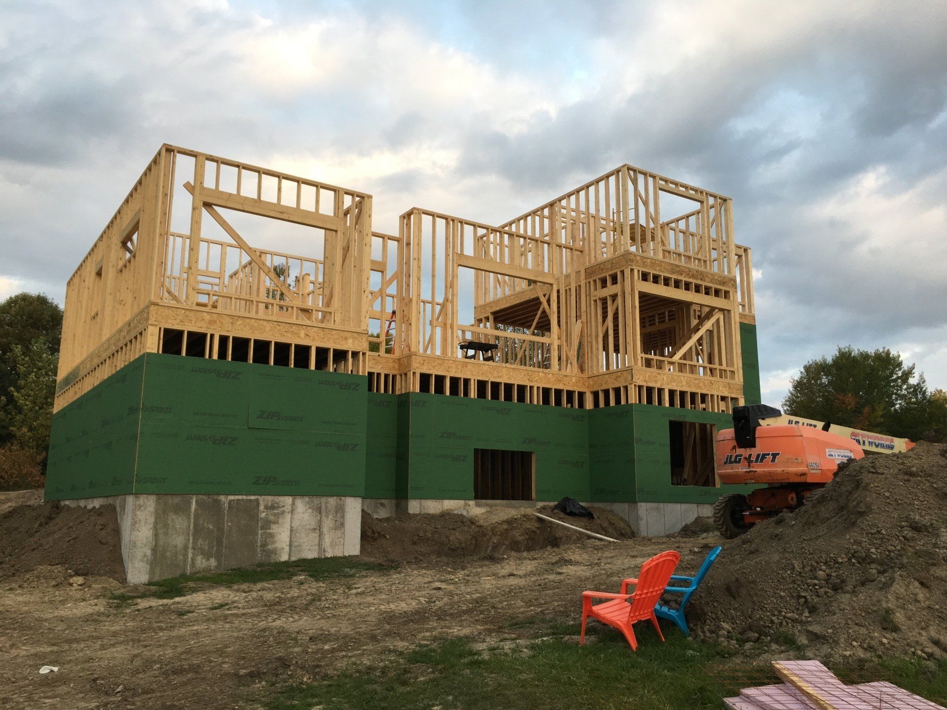 Partially built wooden house on a green foundation with an orange excavator nearby under a cloudy sky
