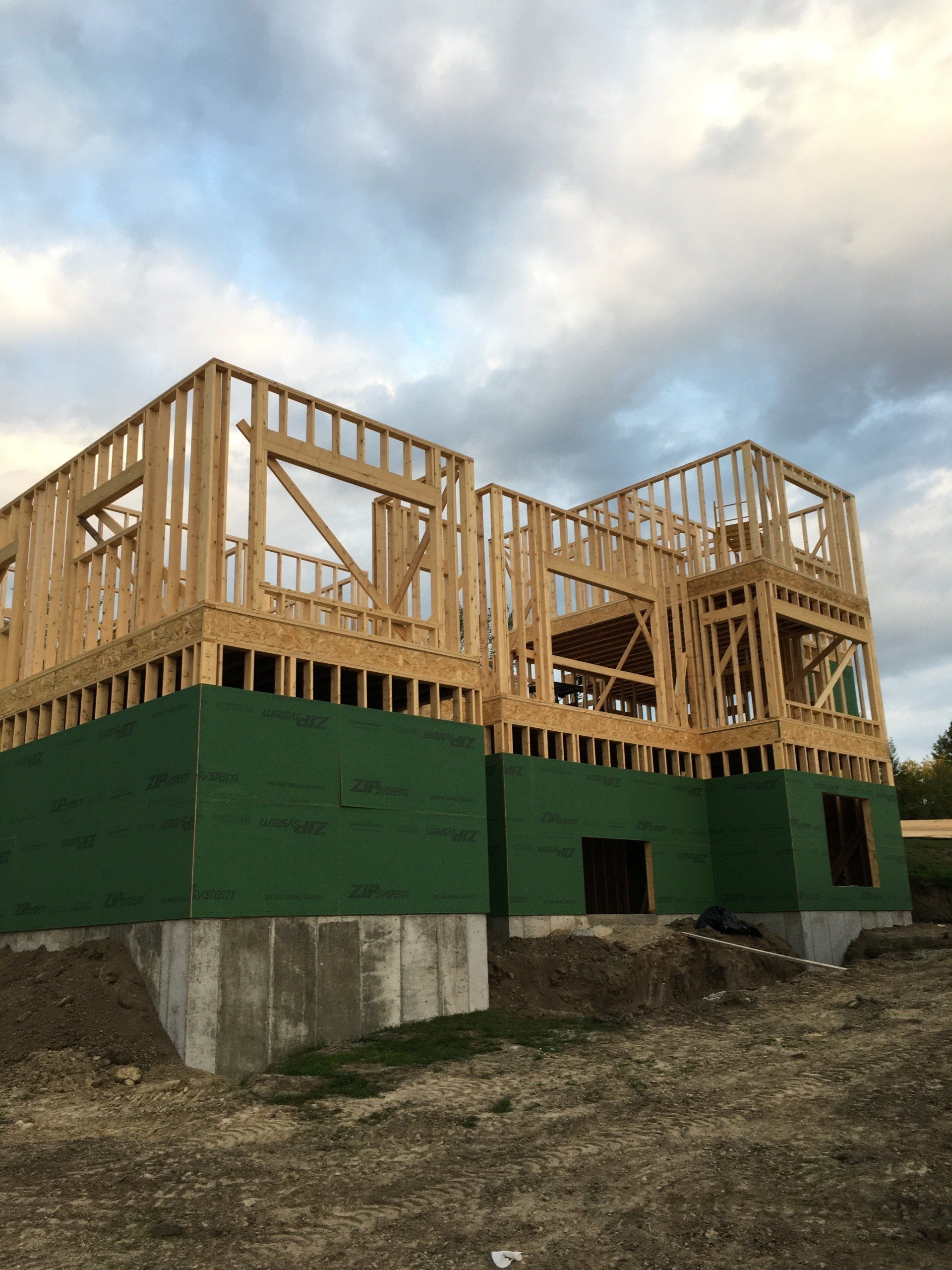 Wood-framed house under construction on a green concrete foundation against a cloudy sky