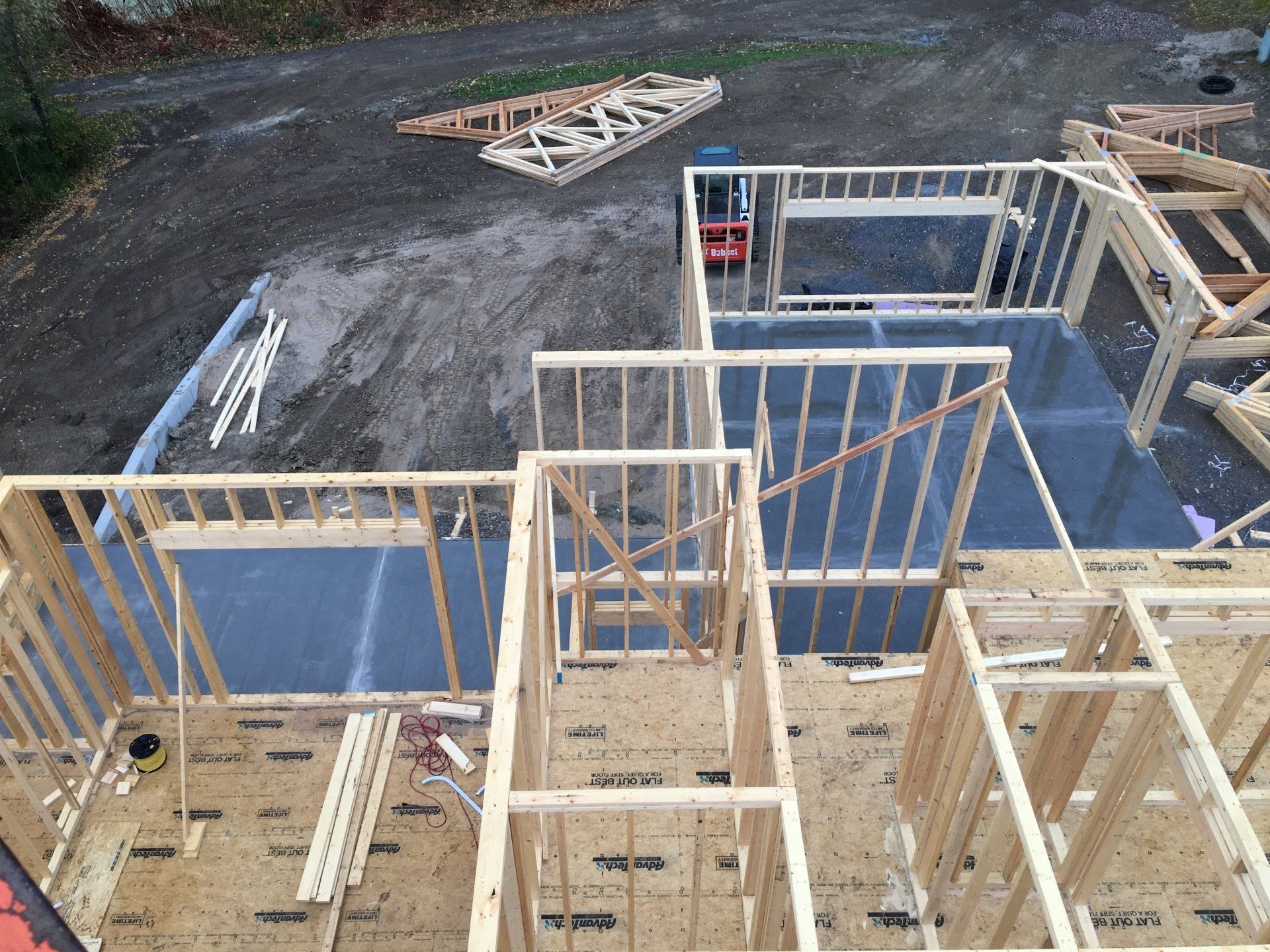 Wood-framed house under construction with exposed walls, stairs, and materials on a gravel lot.