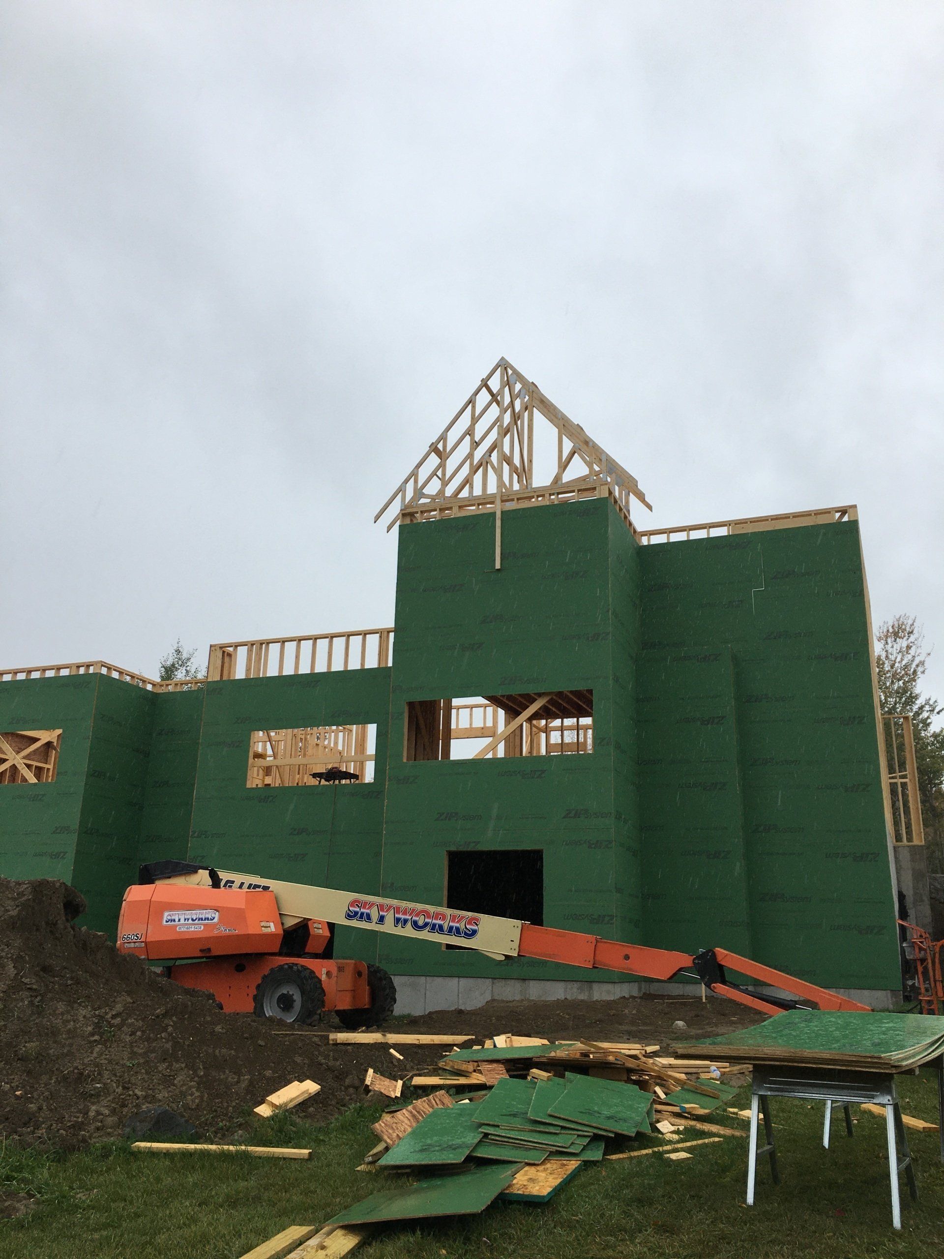 Green-wrapped house under construction with an orange boom lift in front and roof framing above