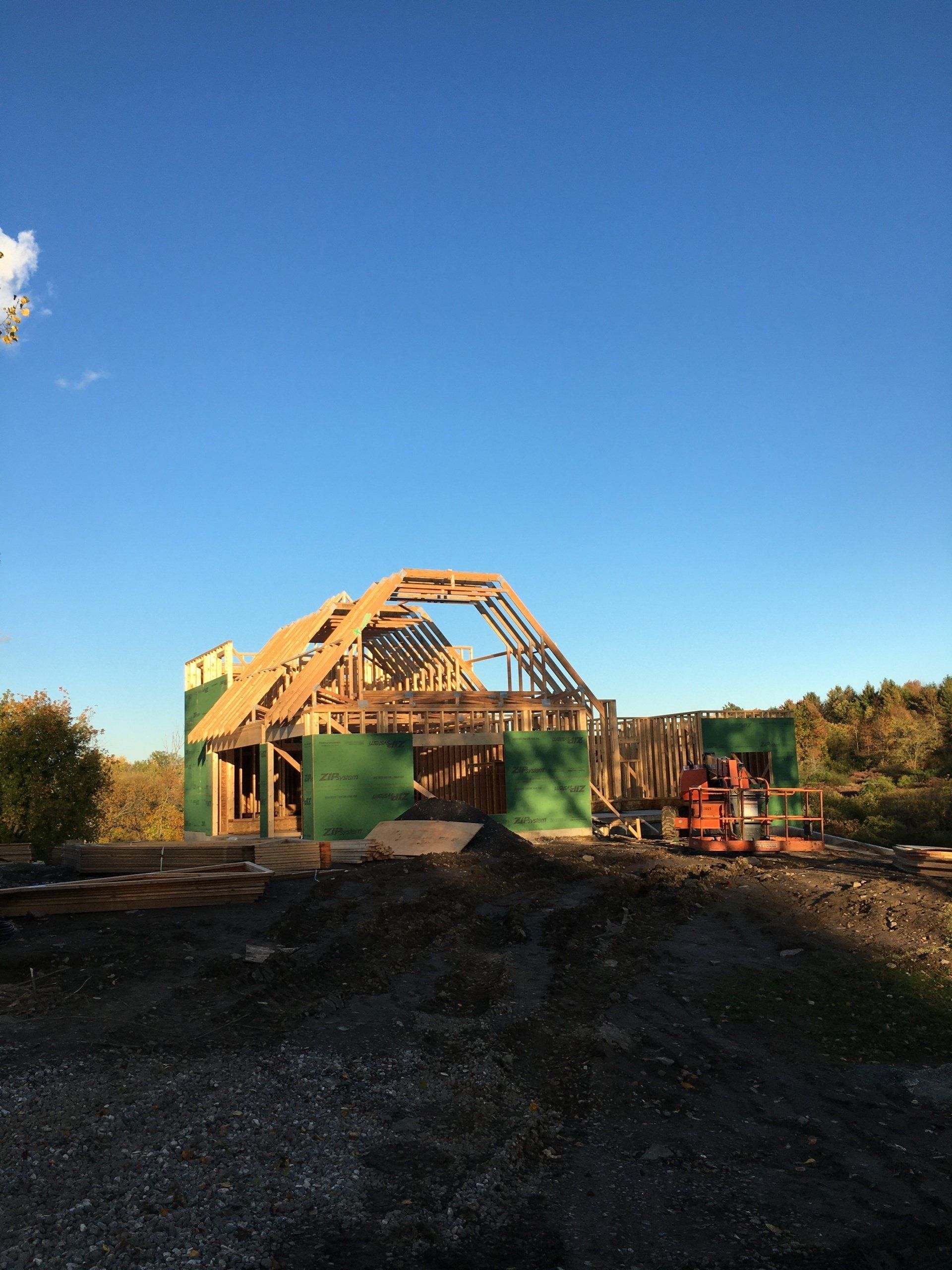 Wooden barn frame under construction on a dirt lot, with a green tank and clear blue sky.