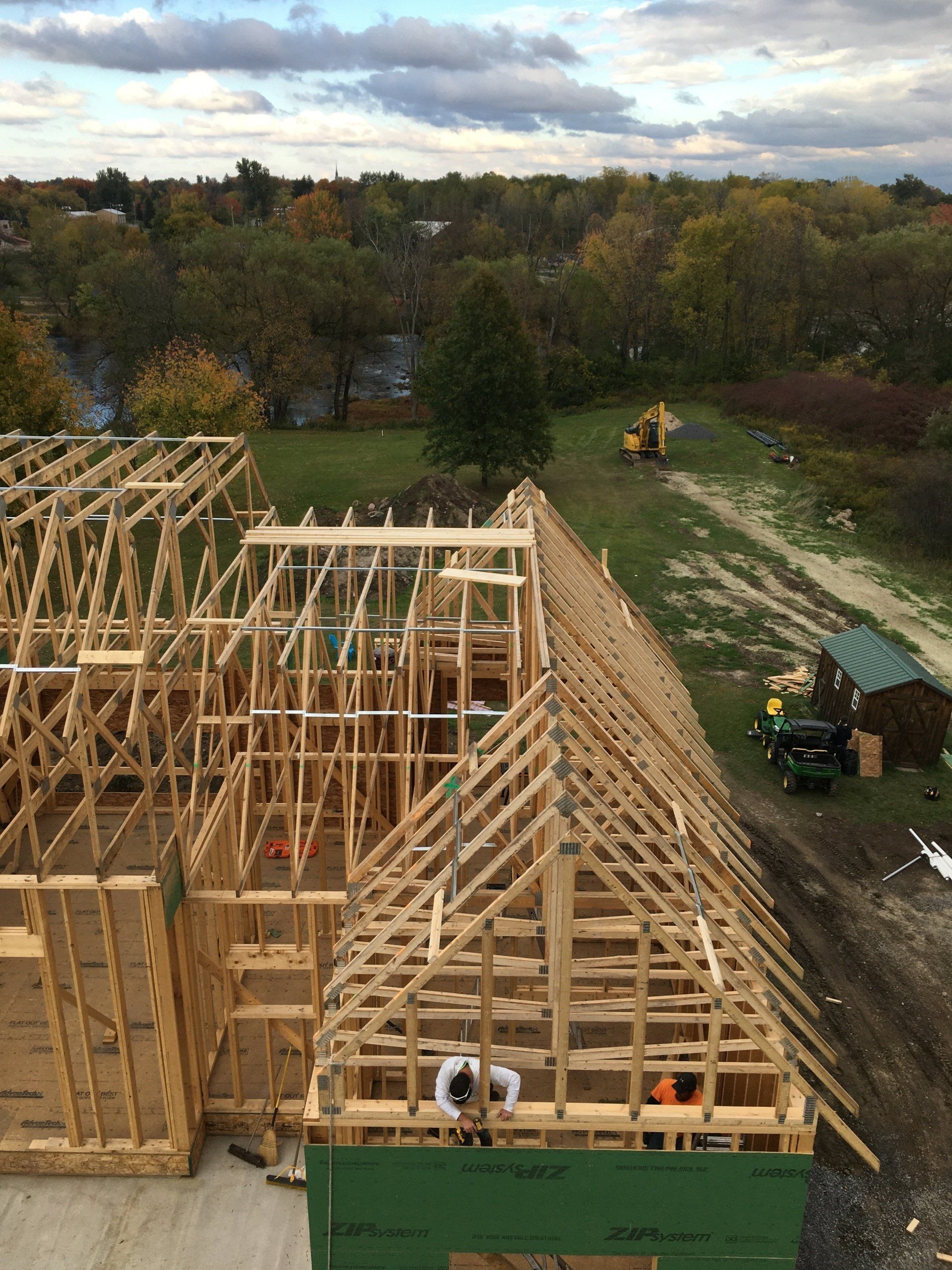 Wood-framed house under construction with rooftop trusses and workers at a rural site under cloudy skies