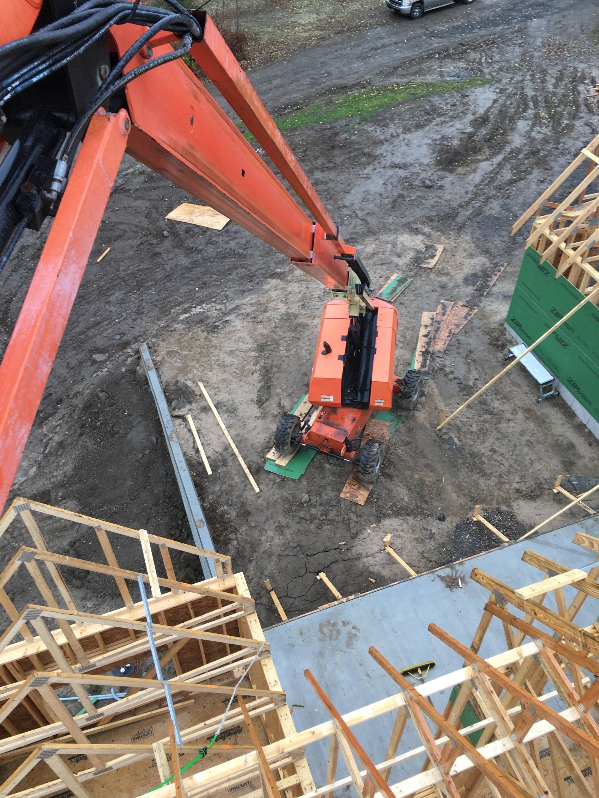 Orange excavator arm over muddy construction site with timber framing and a green dumpster nearby