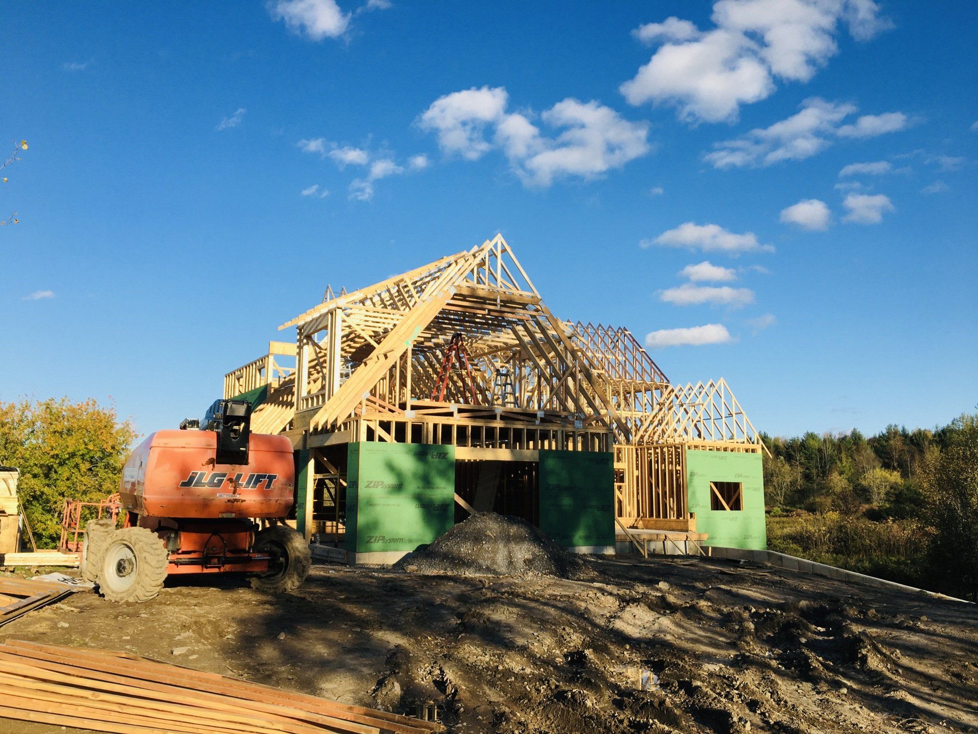 House under construction with exposed wooden framing, a green lift, and an orange excavator on a muddy site.