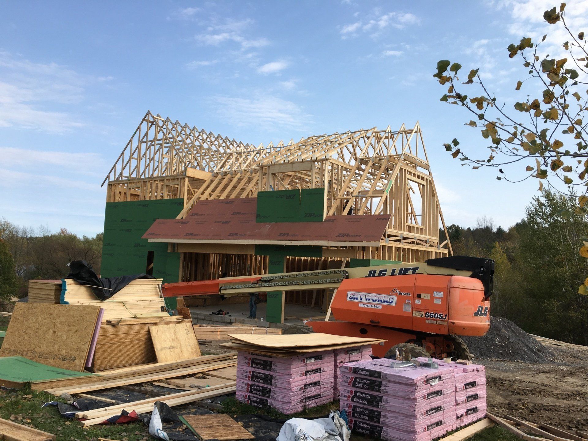 House under construction with wooden roof framing, green sheathing, and construction materials in front