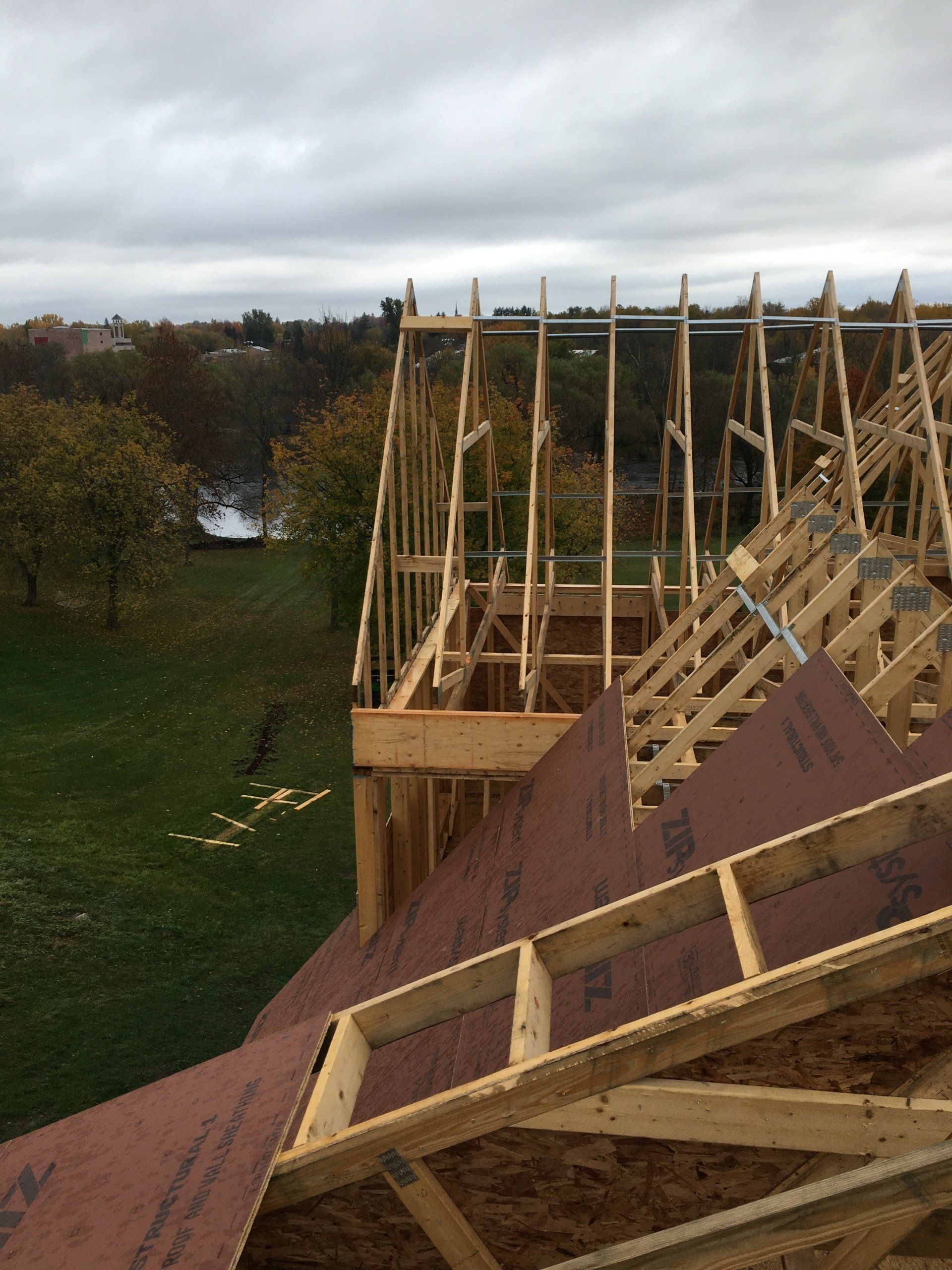 Wooden roof framing under construction beside a grassy field, with brown sheathing and cloudy sky.
