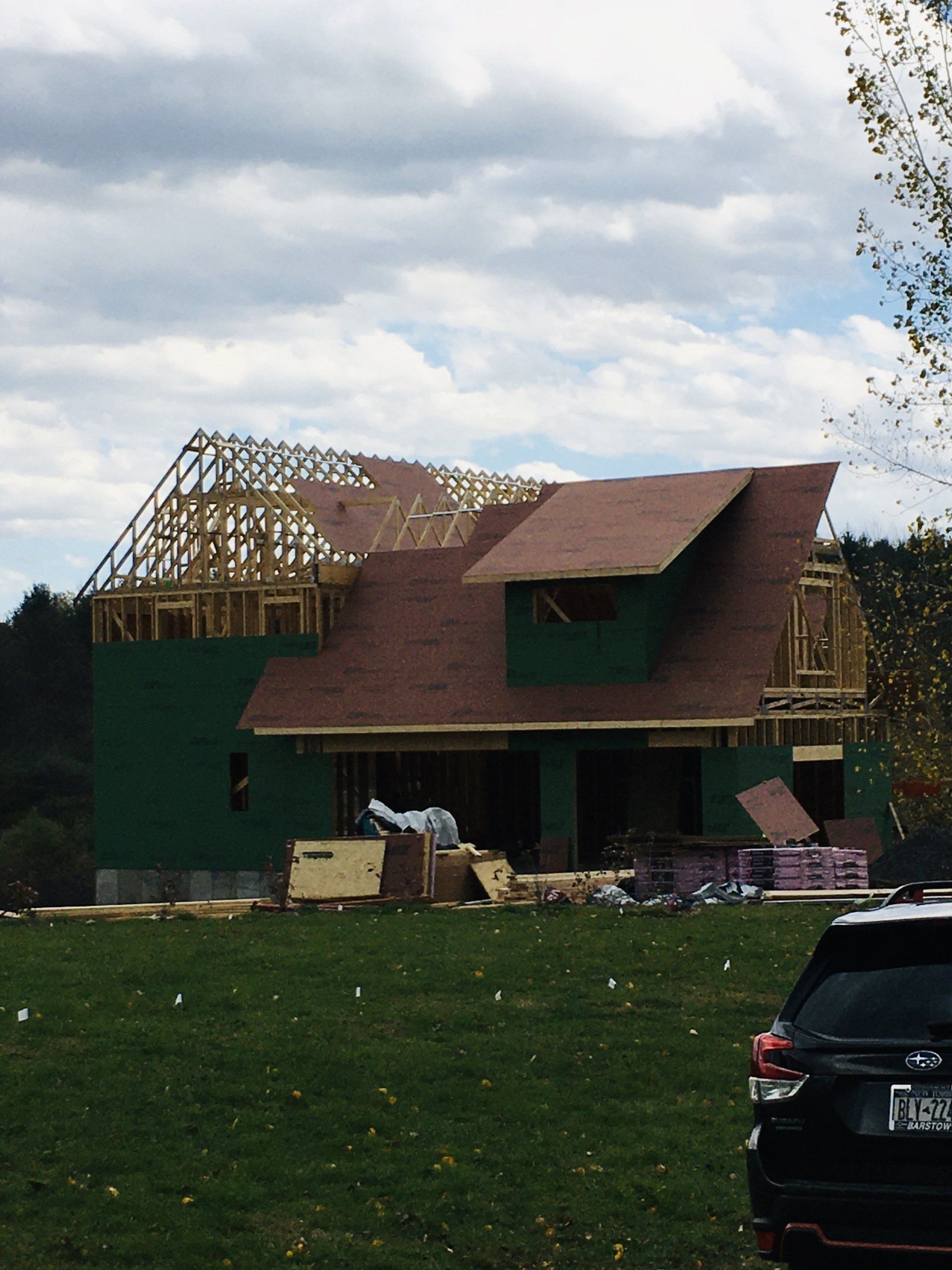 House under construction with exposed roof framing and green exterior, parked car in foreground, cloudy sky