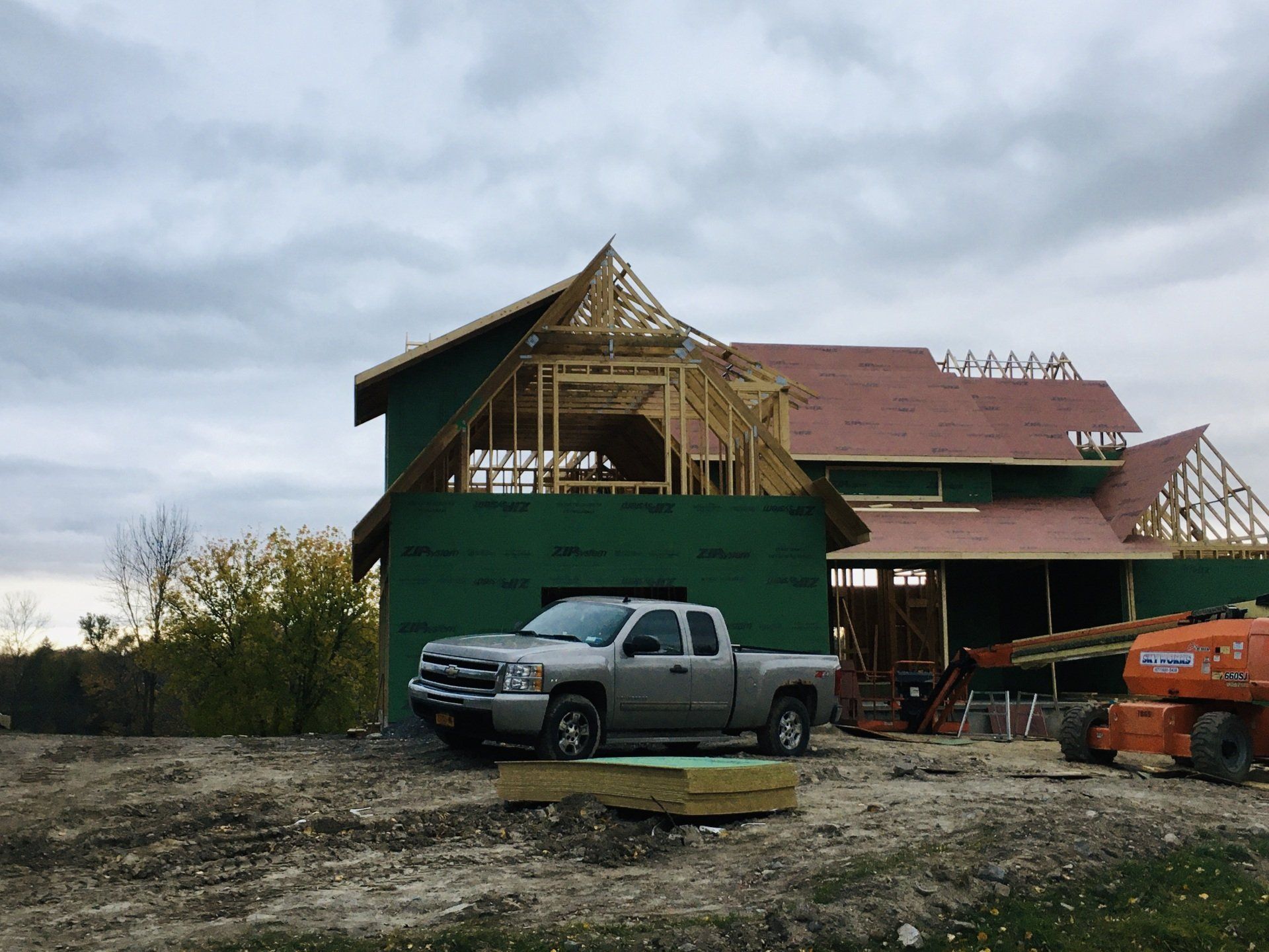 Partially built house with green siding, exposed roof framing, and parked trucks on muddy ground