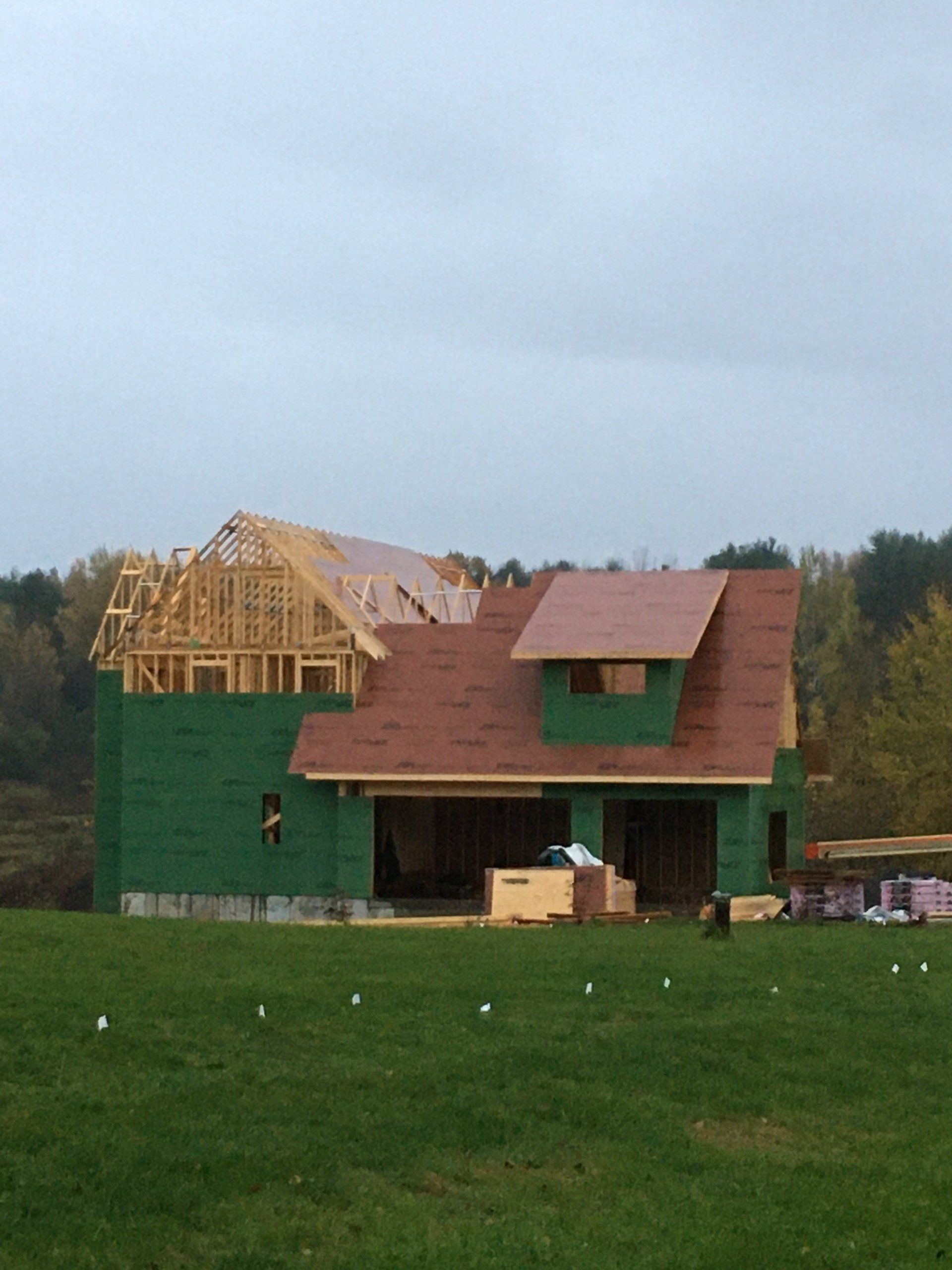 Partially built house with green siding and red roof in a grassy field under cloudy sky