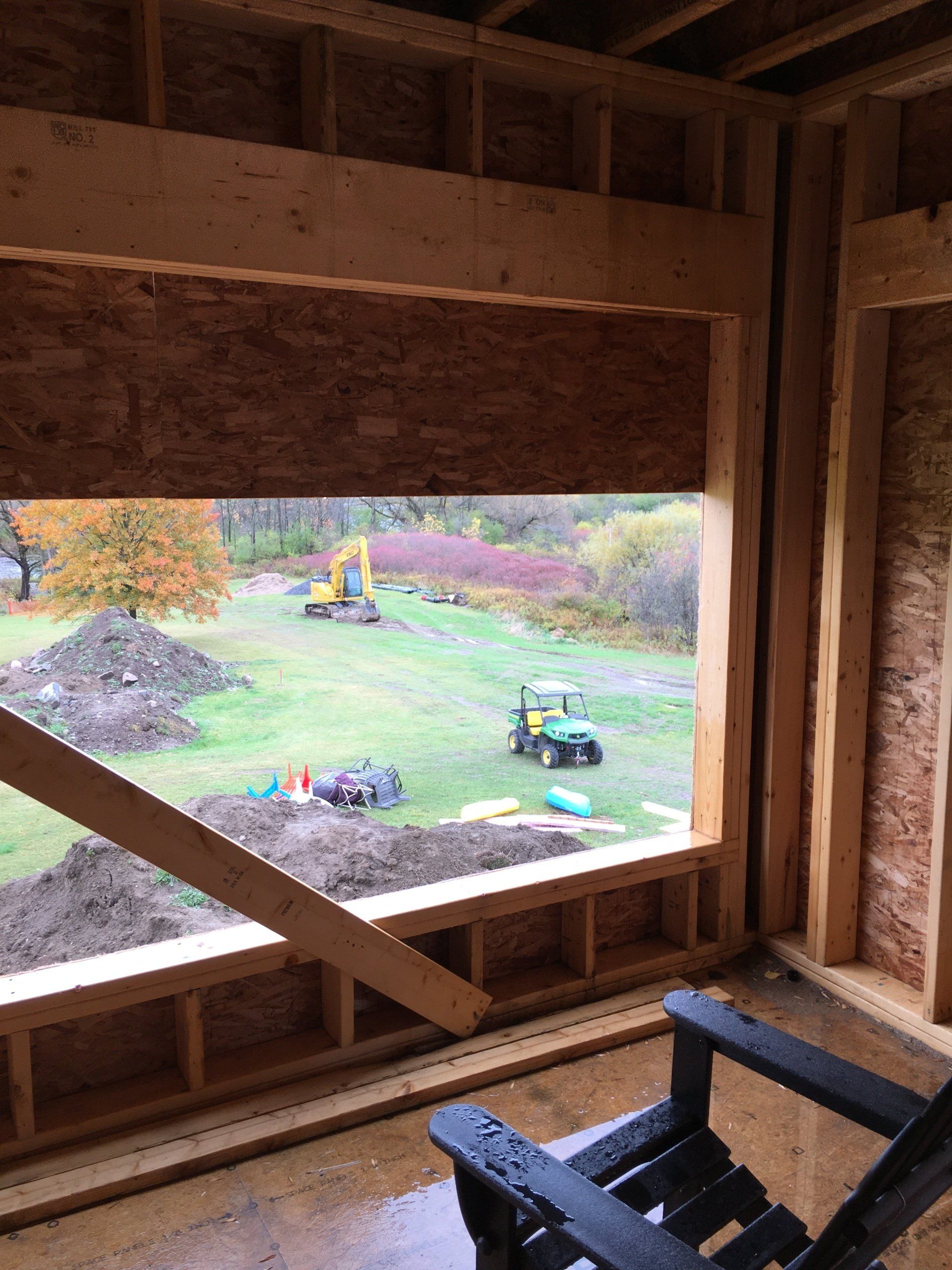 Inside a wooden structure, a large window frames a grassy yard with construction equipment and autumn trees.