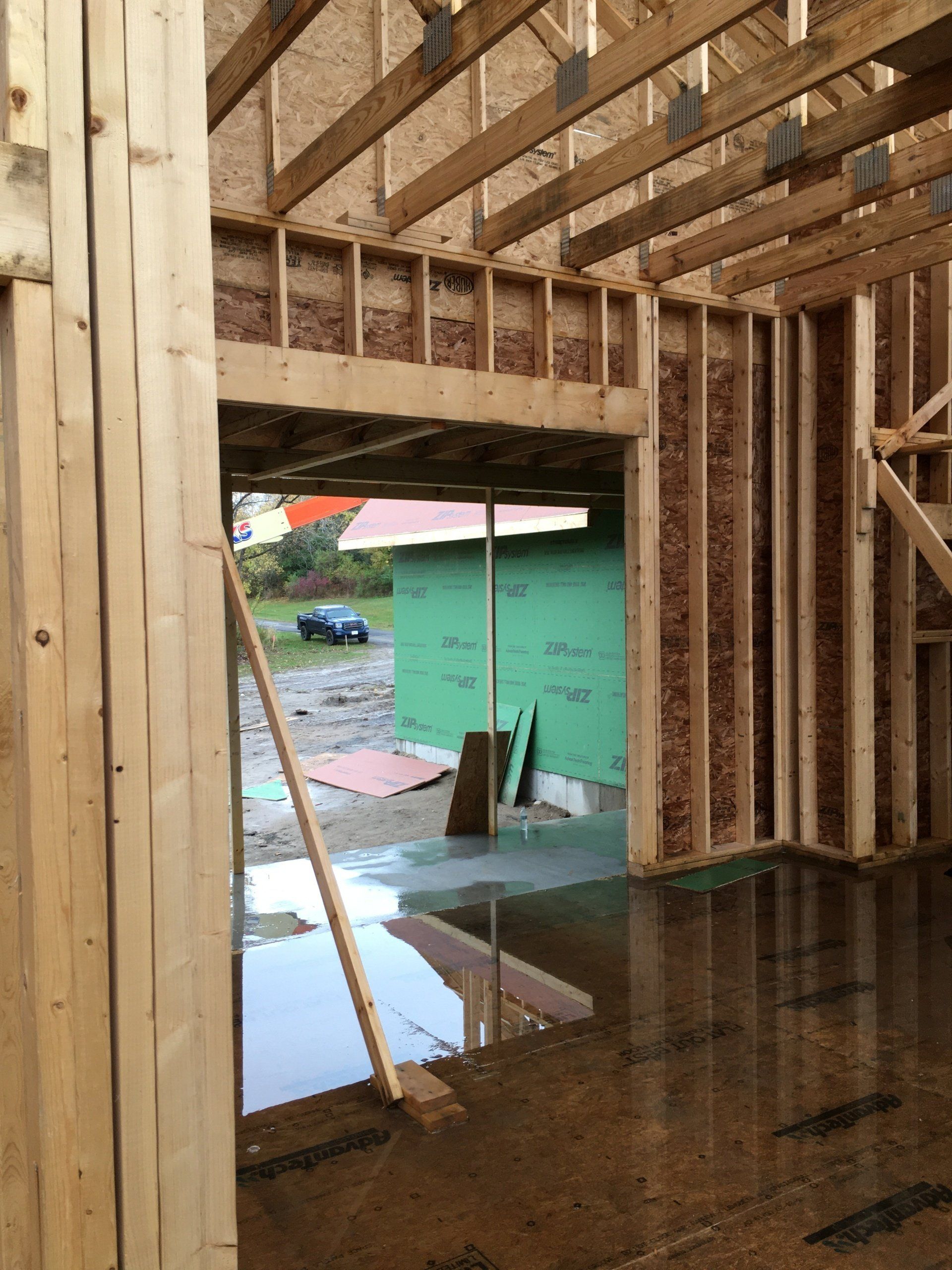 Unfinished wooden interior framing with an open doorway, showing wet floor and a green structure outside.
