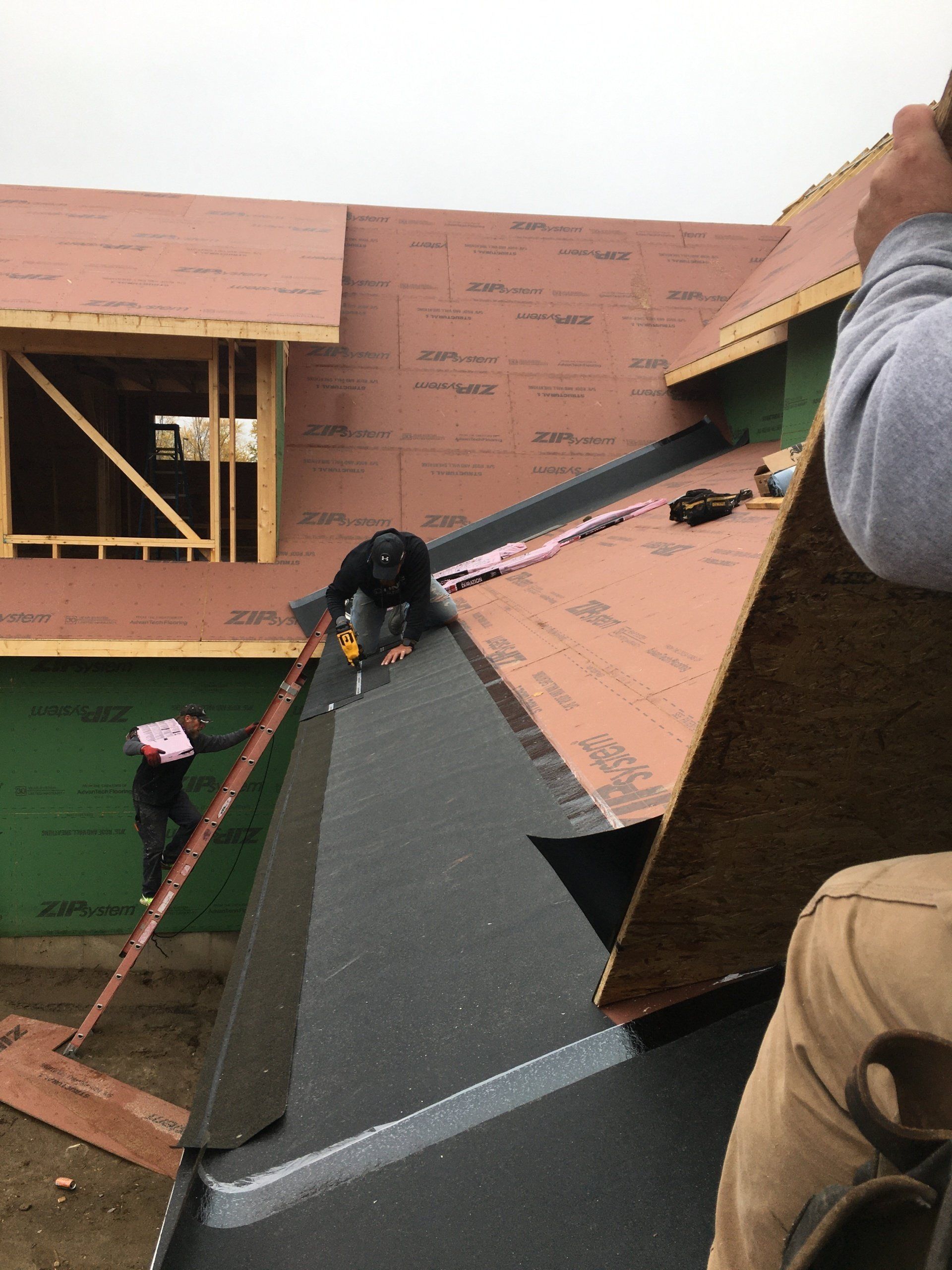 Roofing workers installing underlayment on a steep house roof with a ladder and plywood walls nearby