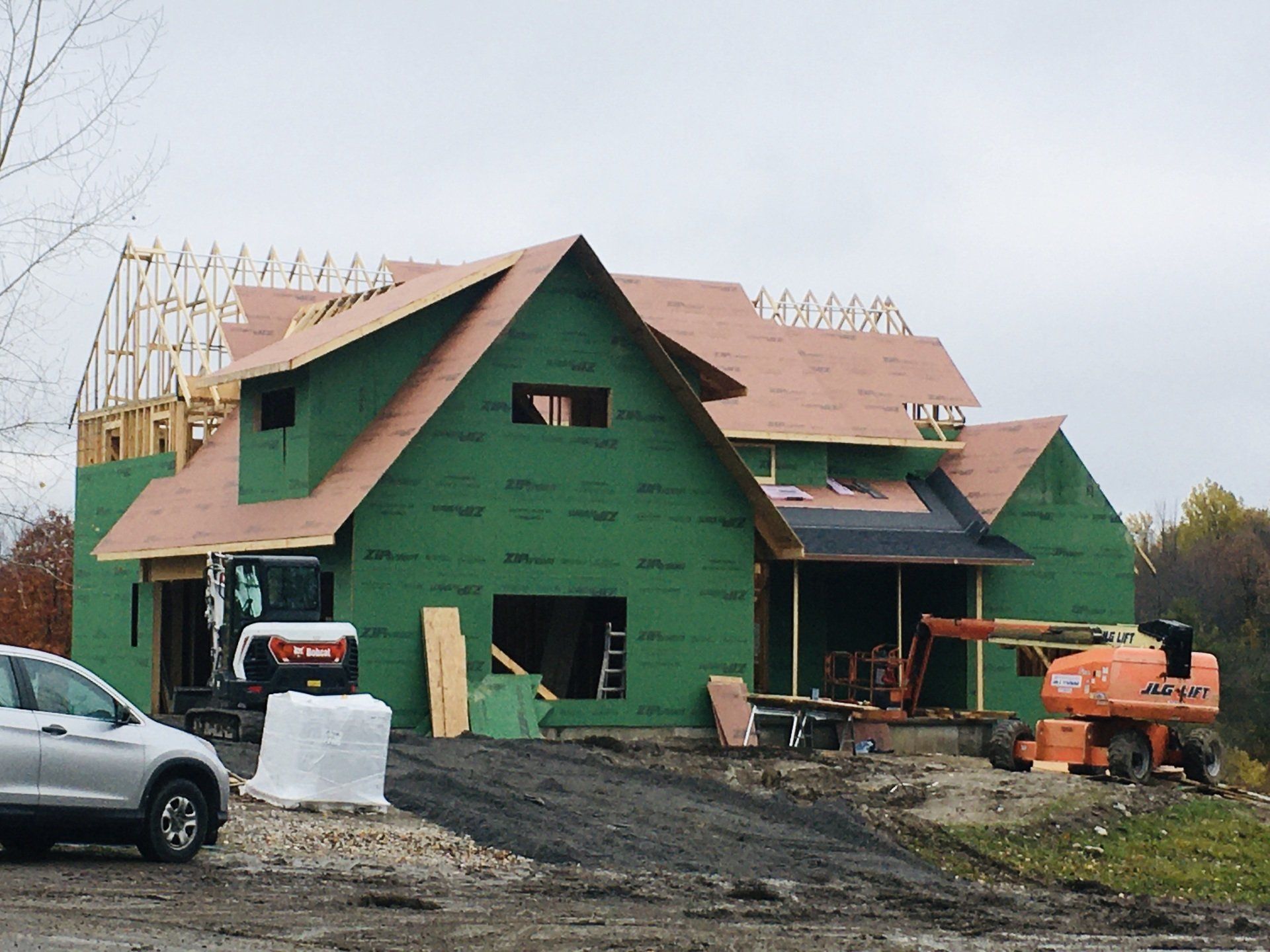 Green house under construction with exposed framing, roof trusses, vehicles, and muddy driveway