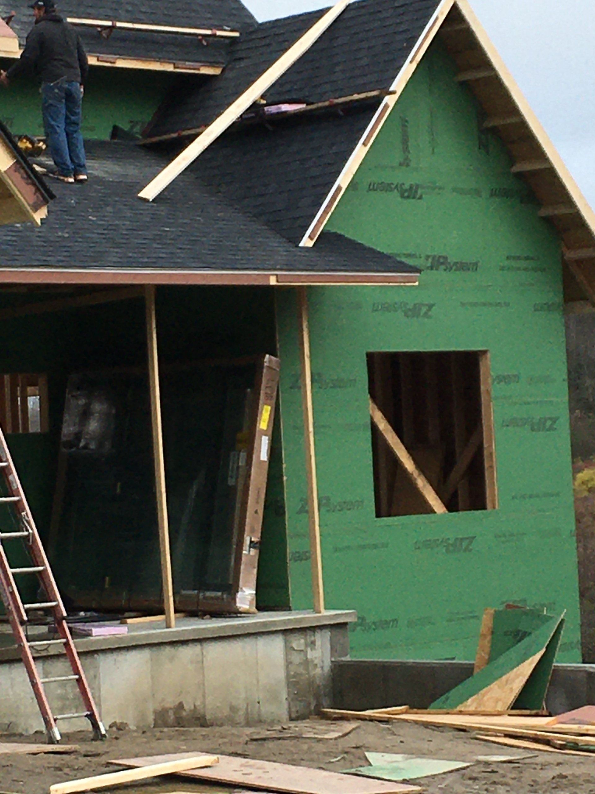 House under construction with green sheathing, roof shingles, and a worker on the roof.