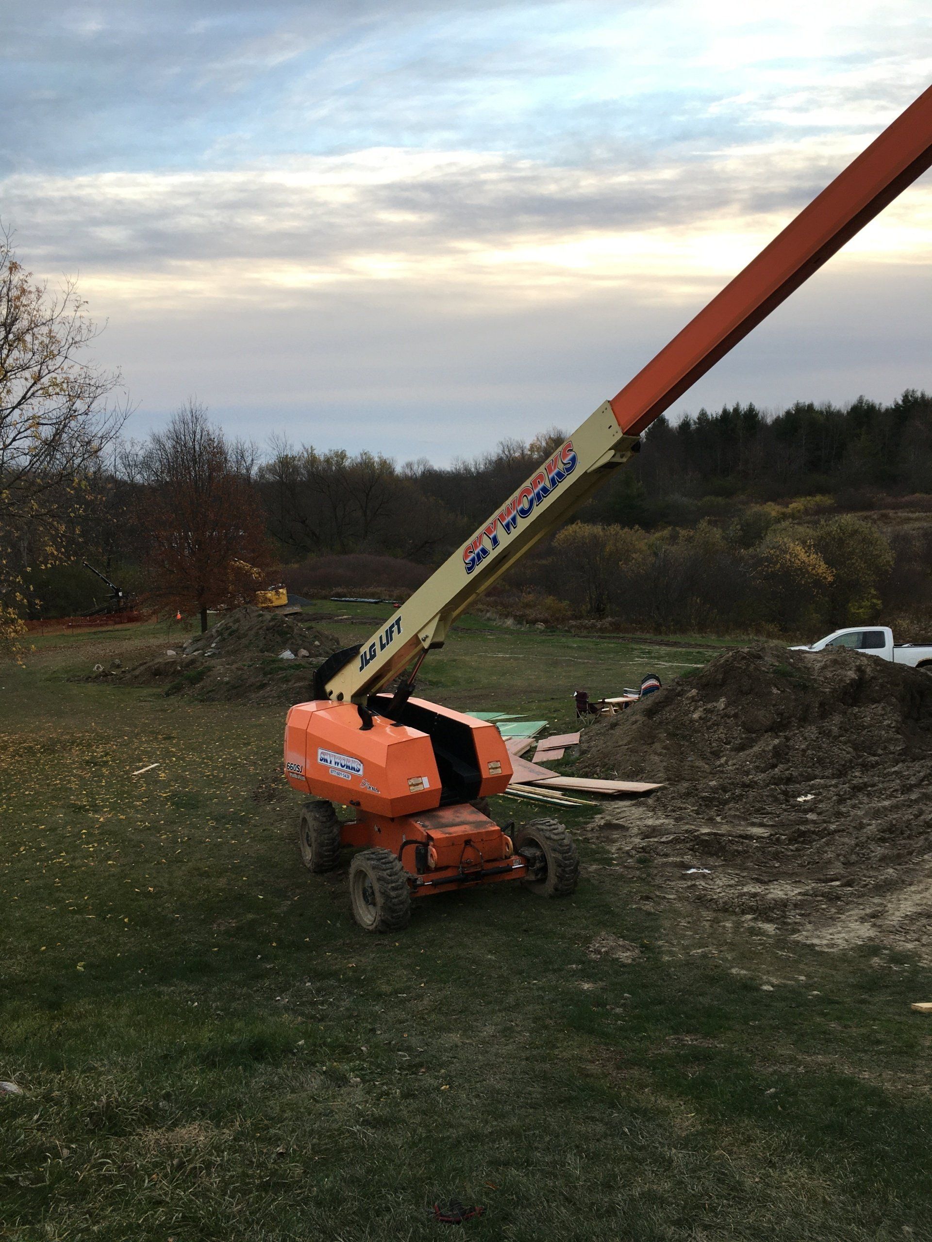 Orange boom lift parked on grassy dirt field at sunset, with arm raised diagonally against cloudy sky