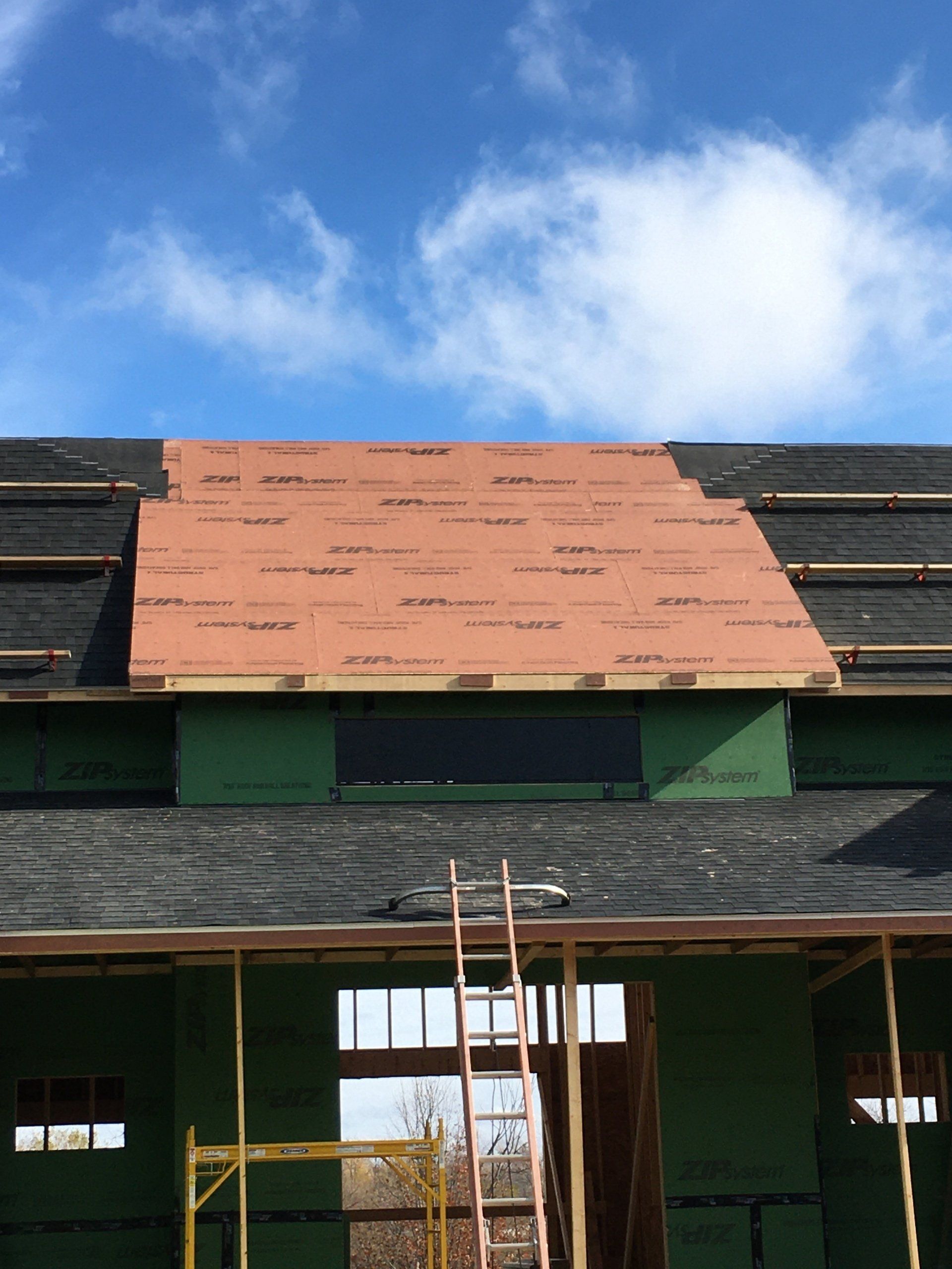 Roof under construction with orange sheathing, ladder, and green siding against a blue sky