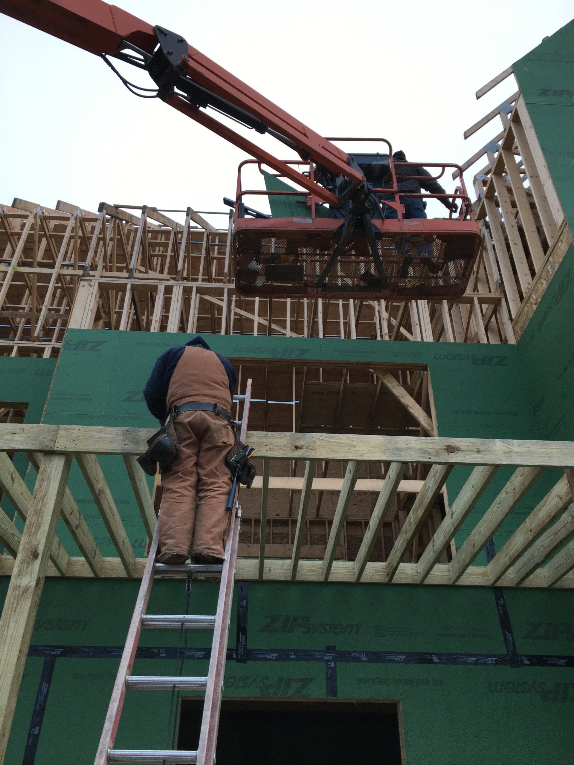 Worker on ladder beneath a crane lifting wooden framing at a construction site