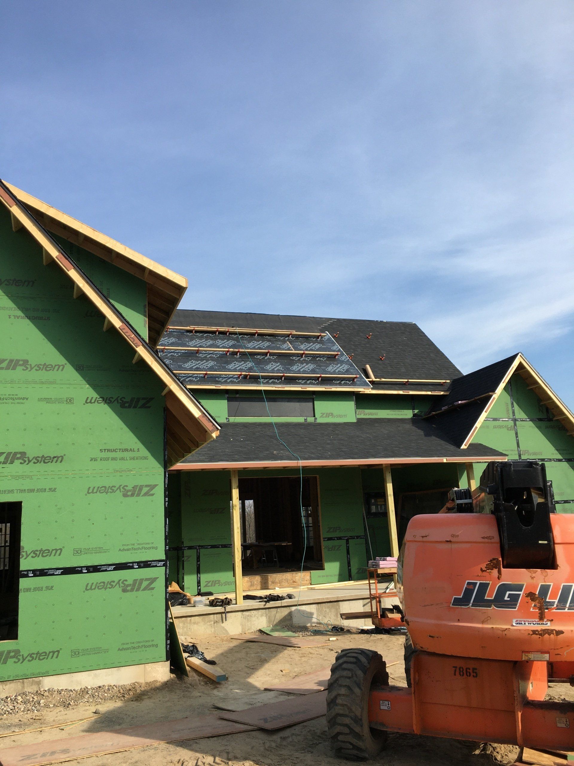 Partially built house wrapped in green sheathing, with a roof under construction and an orange lift in front.